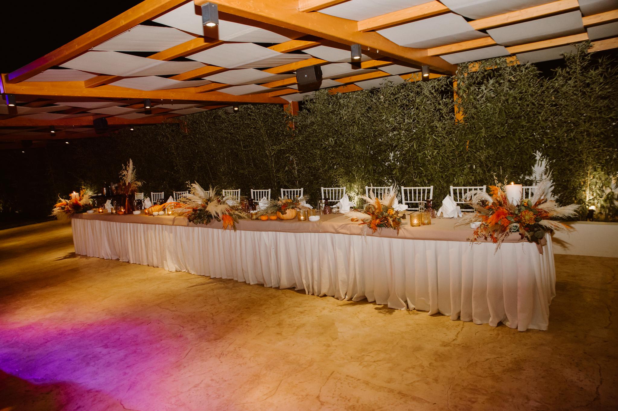 Φωτογραφία κεντρικού τραπεζιού κάτω από την ξύλινη πέργκολα η οποία έχει πανιά από επάνω. Στο κεντρικό τραπέζι βρίσκετε στολισμός που αποτελείτε από Pampas μπεζ και φούξια μαζί με λευκά και φούξια τριαντάφυλλα και πρασινάδες. Στο βάθος υπάρχει η πρασινάδα που φαίνεται από το κτήμα.