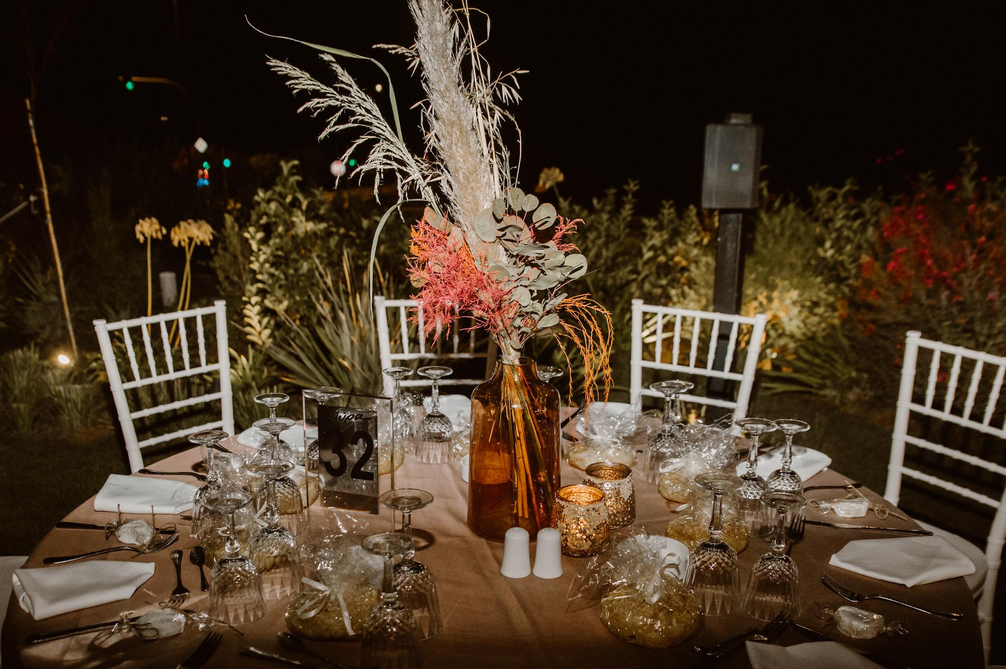 Κοντινή φωτογραφία από το τραπέζι που έχει στήσει το catering με γυάλινα ποτήρια και μαχαιροπίρουνα. Στο κέντρο έχει τον στολισμό ο οποίος αποτελείτε από pampas mpez και φούξια και πρασινάδες