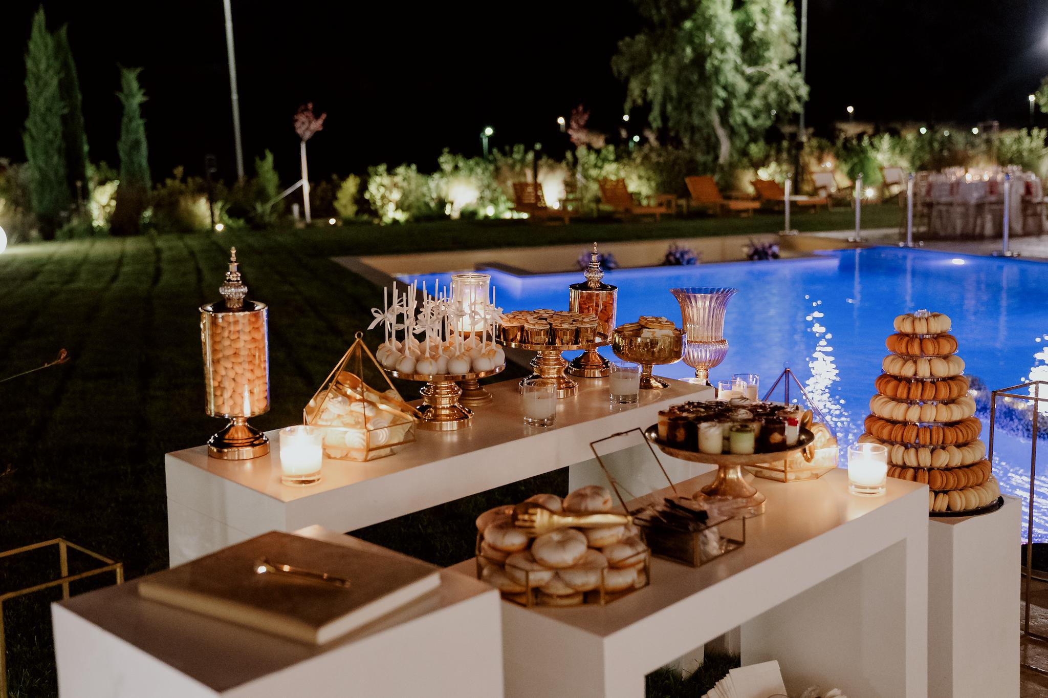 candy bar στην είσοδο του κτηρίου σε λευκά τραπέζια επάνω. Επάνω στα τραπέζια έχει τουρτιέρα με γλυκά και μάκαρονς . Χρυσές βάσεις με ντόνατς και κουφέτα σε βάζα . Τα καπκεικ είναι επάνω σε χρυσή τουρτιέρα