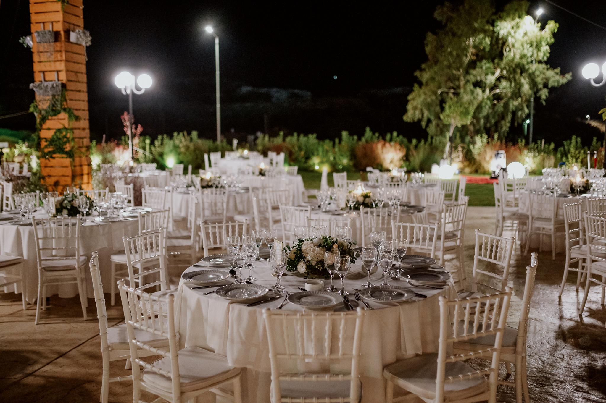 νυχτερινή λήψη φωτογραφίας με αναμμένα τα φώτα. Τραπέζια cateirng με ποτήρια και πιάτα επάνω στολισμό με τριαντάφυλλα λευκά και σάπιο μήλο με φόντο πράσινο γκαζόν και πολύχρωμα λουλούδια.