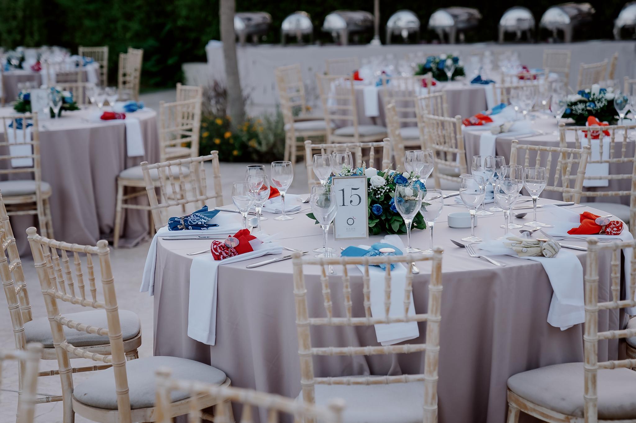 Κοντινή φωτογραφία στα τραπέζια του κτήματος , λουλούδια βρίσκονται επάνω στα τραπέζια τα οποία περιέχουν μπλε τριαντάφυλλα και λευκά όπως και γυψοφύλλι . Επάνω στο κάθε τραπέζι βρίσκονται τοποθετημένες και η μπομπονιέρες. Στο βάθος φαίνεται το γκαζόν όπως επίσης και και ο πράσινος κήπος με τα λουλούδια.