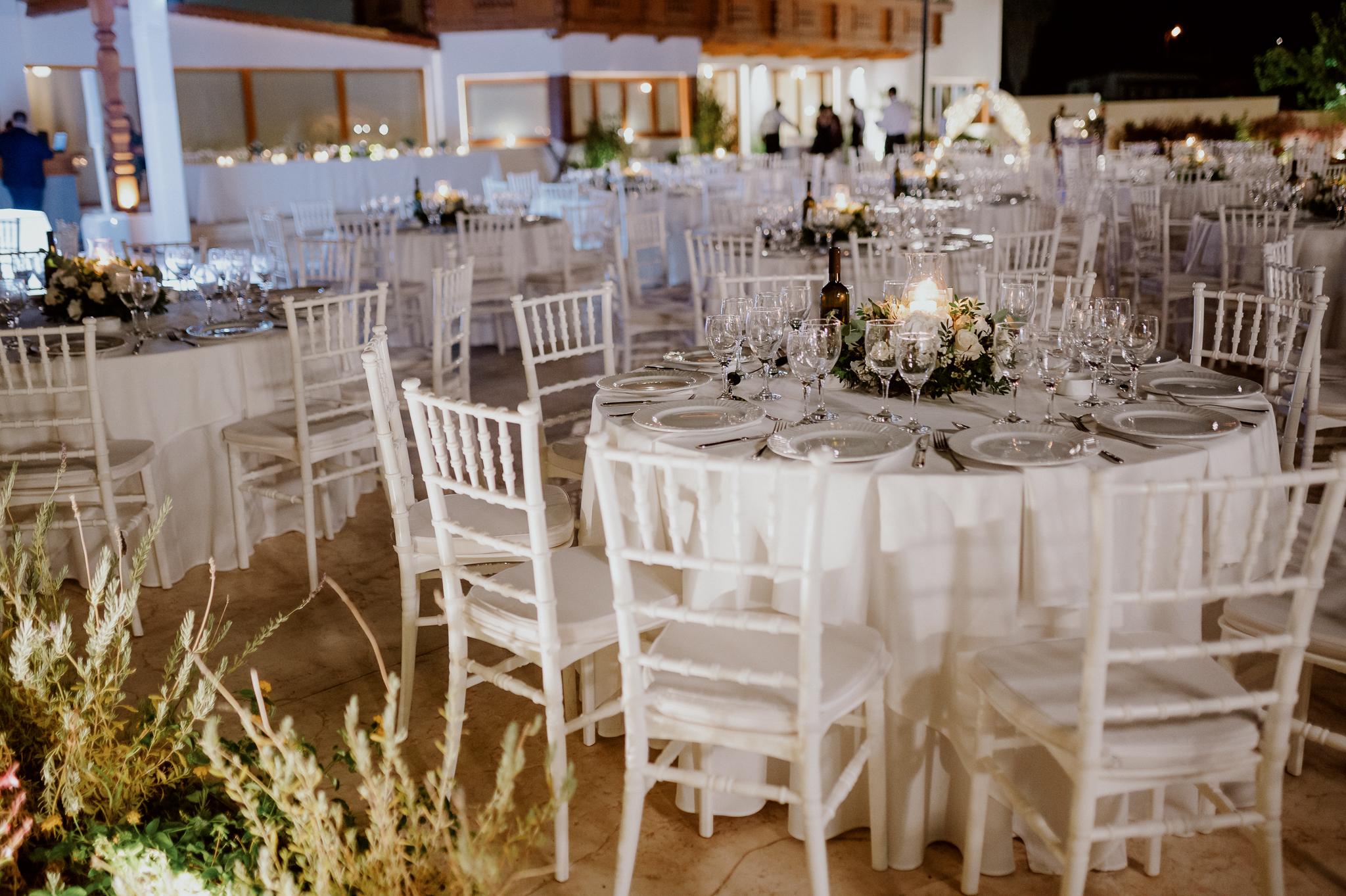 Ροτόντες στρωμένες με λευκά τραπεζομάντηλα στον εξωτερικό χώρο κτήματος. Επάνω τα τραπέζια έχουν στολισμό με λευκά τριαντάφυλλα και κερί στην μέση. Η καρέκλες είναι λευκές tiffany και επάνω στο κάθε τραπέζι έχει ένα μπουκάλι κρασί