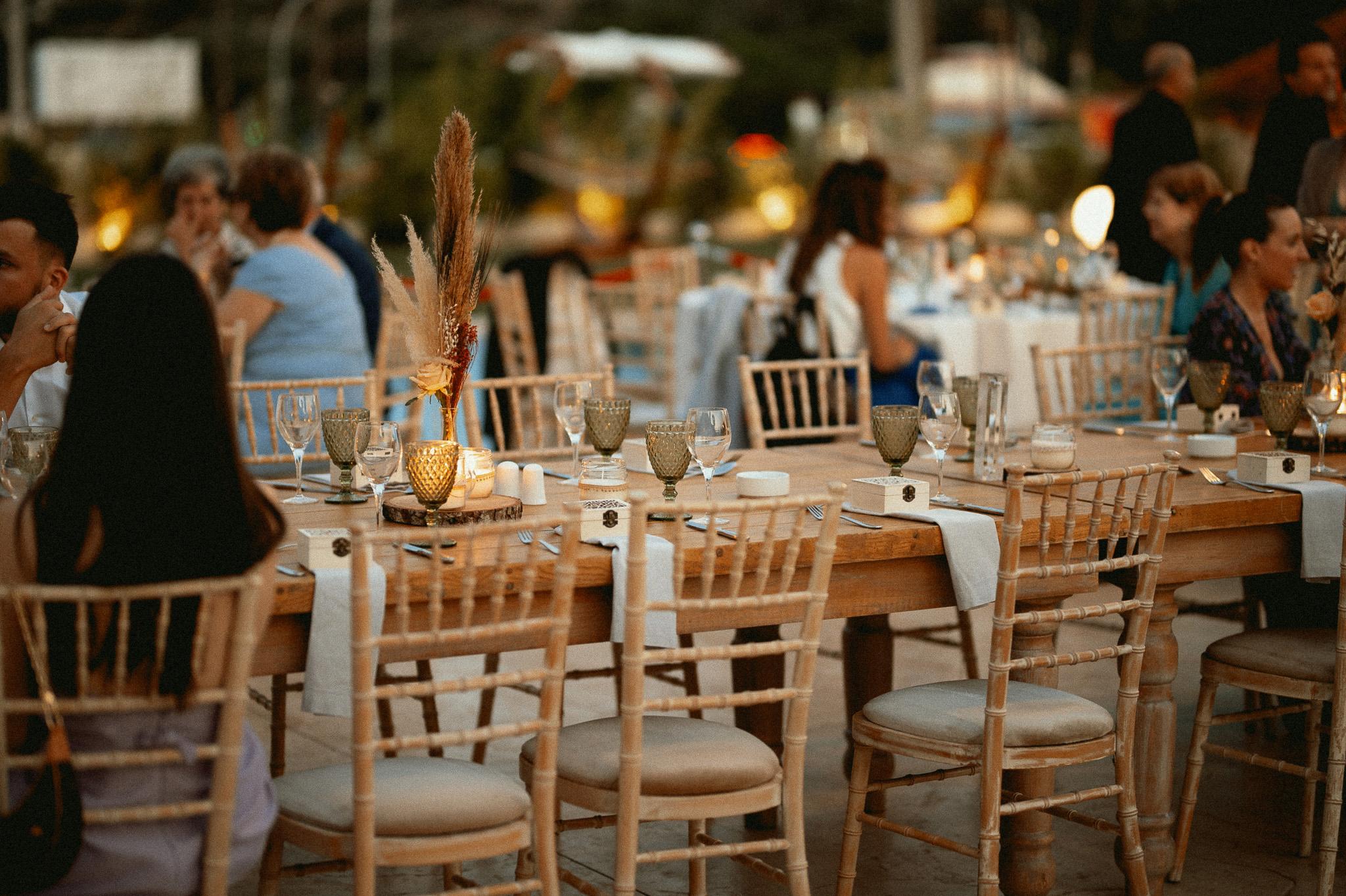 Μοναστηριακό ξύλινο τραπέζι με καρέκλες tiffany. Άνθρωποι κάθονται στο μοναστηριακό τραπέζι . Επάνω στο μοναστηριακό τραπέζι βρίσκονται τοποθετημένα ποτήρια και μαχαιροπίρουνα.