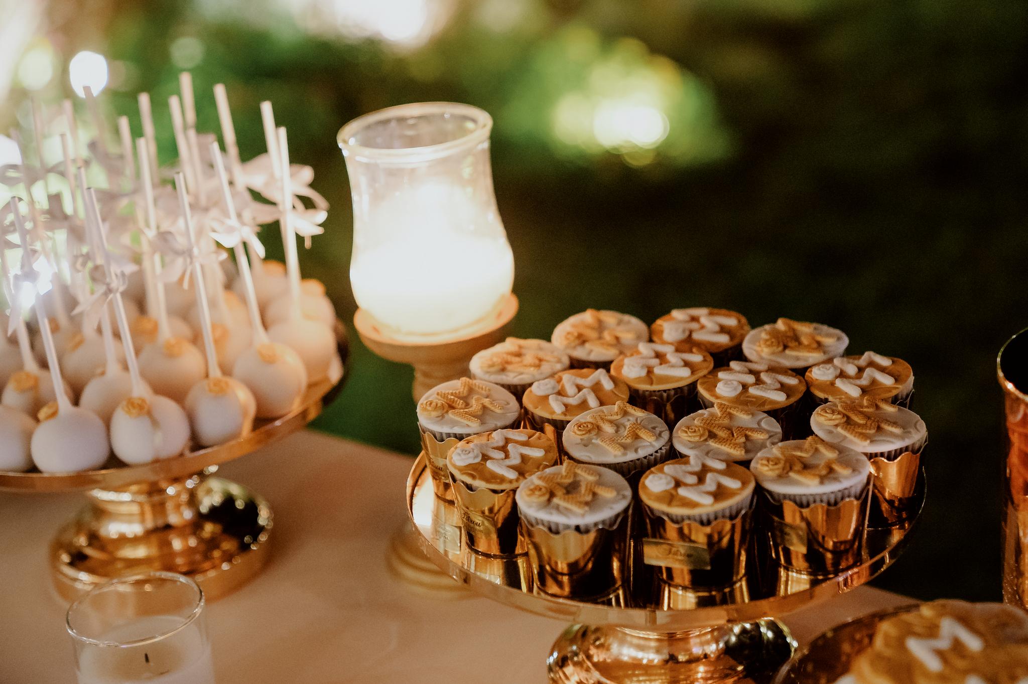 candy bar κοντινή λήψη σε γλυκά τα οποία είναι cup cake και macaron με τα αρχικά του ζευγαριού από πάνω σε ζαχαρόπαστα σε χρώματα λευκά ροζ και χρυσά . Επάνω στο τραπέζι έχει cake pops όπως επίσης και στολισμό ο οποίος είναι λουλούδια λευκά τριαντάφυλλα