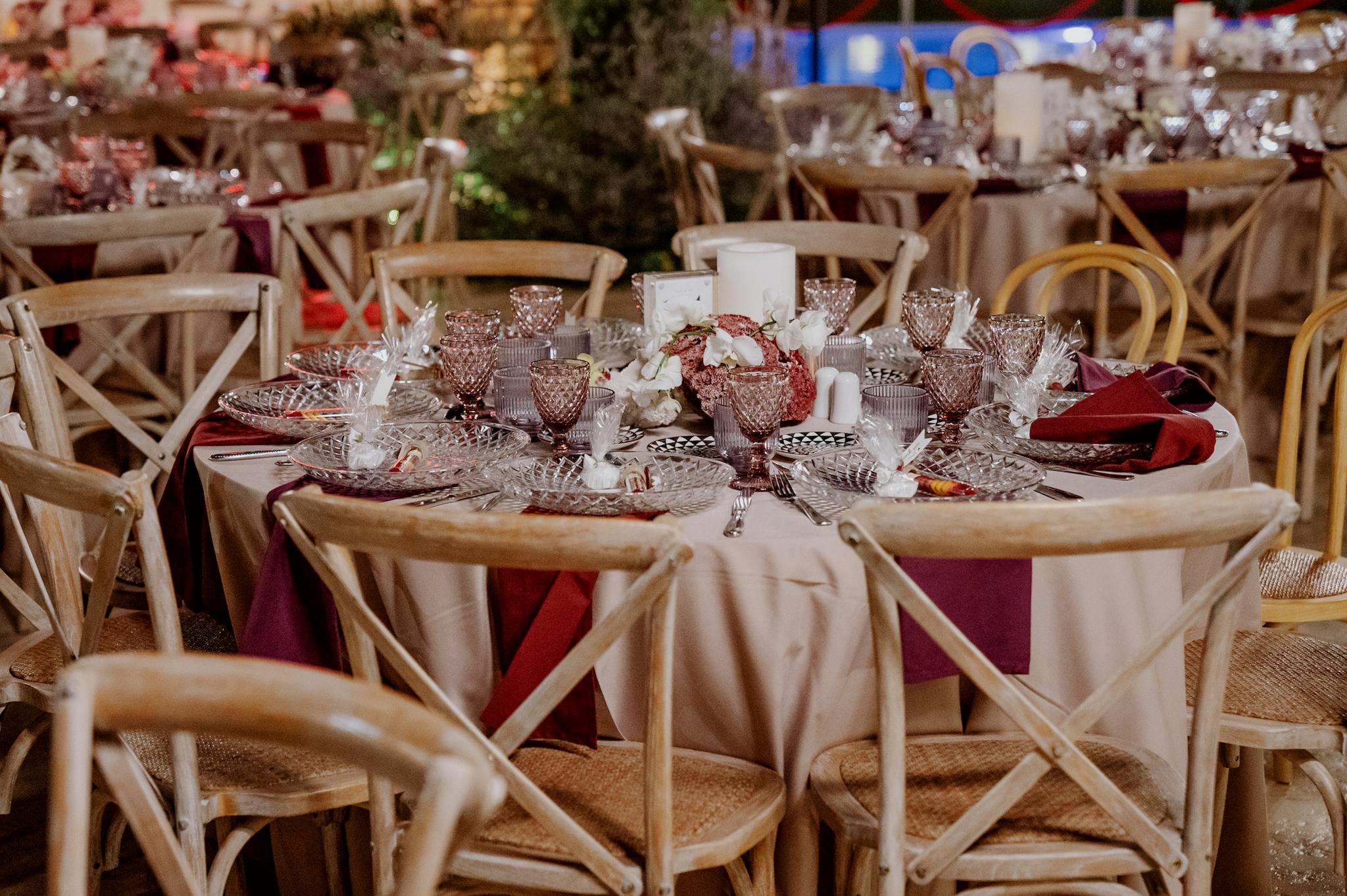 Κοντινή φωτογραφία με ροτόντες τραπέζια περιμετρικά από τα τραπέζια είναι ξύλινες καρέκλες. Τα τραπέζια έχουν μπεζ τραπεζομάντηλα και επάνω μπορντό ατομικές πετσέτες. Στο βάθος είναι το αυτοκινούμενο βαν μπαρ το οποίο το χρησιμοποιούν οι καλεσμένοι για να παίρνουν ποτό . Στο βάθος φαίνεται το γκαζόν αλλά και τα φυτά που έχει το κτήμα. Στο βάθος φαίνεται το πράσινο και ο κήπος