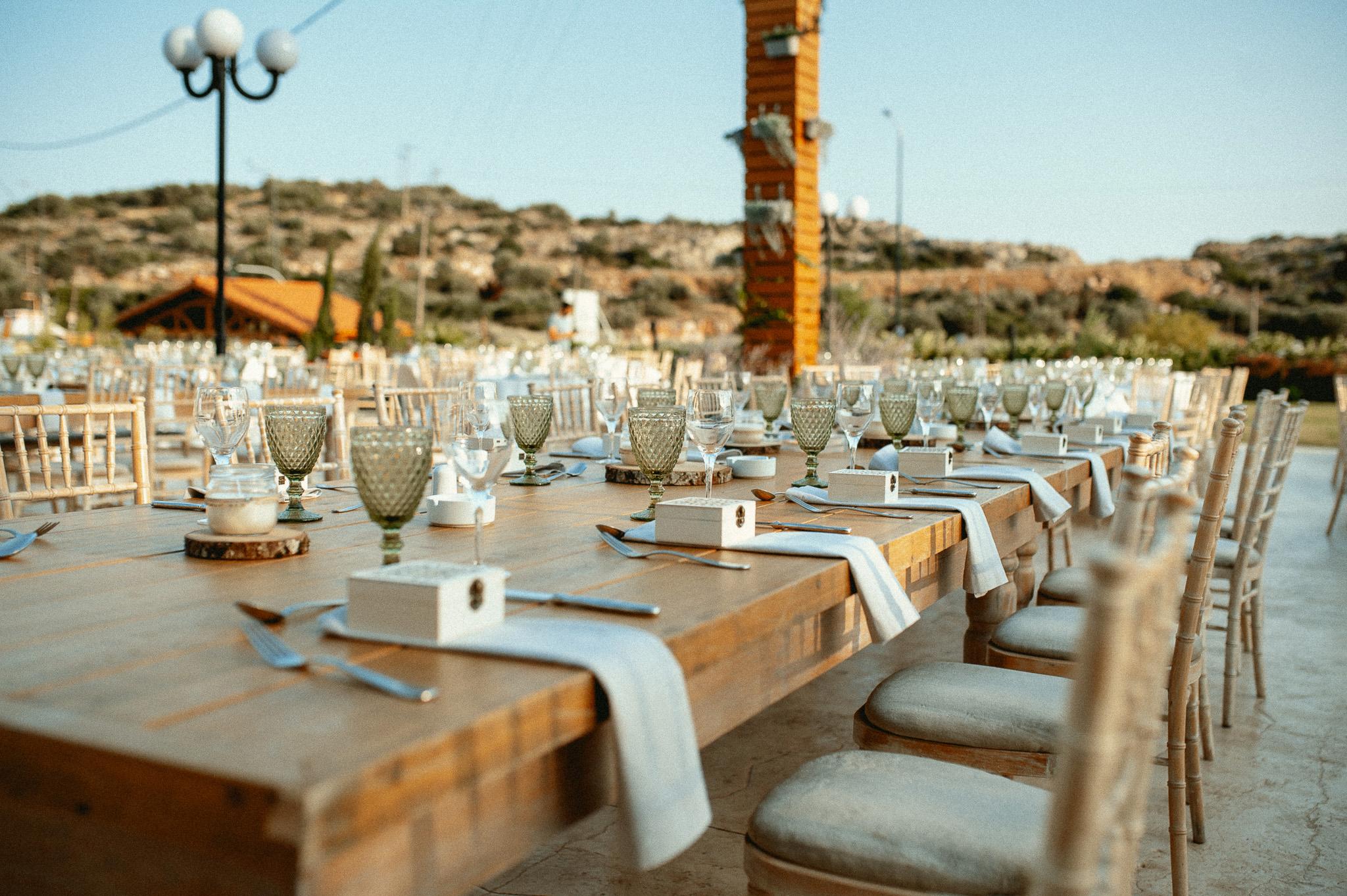 Μοναστηριακό τραπέζι με πράσινα ποτήρια και λευκές πετσέτες ατομικές με τις μπομπονιέρες επάνω στις πετσέτες και καρέκλες ξύλινες. Επάνω στα τραπέζια το catering έχει στρώση με ποτήρια και μαχαιροπίρουνα όπως επίσης υπάρχει στο κάθε άτομα και μια μπομπονιέρα . Στην μέση του τραπεζιού υπάρχει ένα ξύλινος κορμός που επάνω που έχει κεράκι και υπάρχει και ένα βάζο το οποίο μέσα έχει pampas σε χρώματα μπεζ και ένα τριαντάφυλλο λευκό