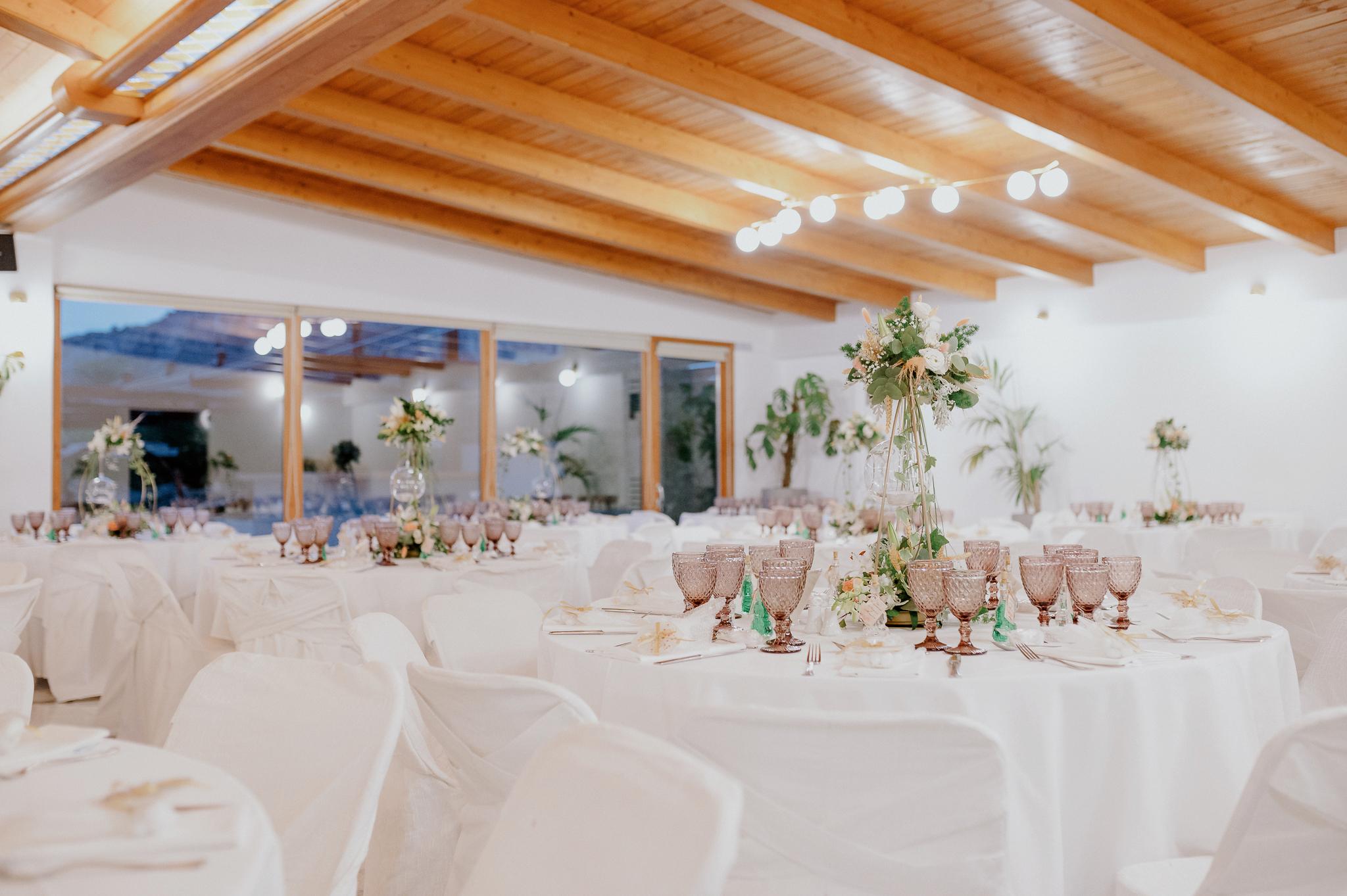 Απομακρυσμένη φωτογραφία στον εσωτερικό χώρο με ροτόντες και τραπέζια περιμετρικά από τις ροτόντες. Επάνω τα τραπέζια είναι στρωμένα με ποτήρια και μαχαιροπίρουνα. Στην μέση των τραπεζιών υπάρχει στολισμός όποιός αποτελείτε από γαρδένιας μαζί με λευκά και σομόν τριαντάφυλλα όπως και πρασινάδες. Στην μέση από το στρογγυλό που αποτελούνται κρέμεται ένα κεράκι . Στο βάθος φαίνονται οι τζαμαρίες του κτήματος που αφήνουν να δεις των έξω χώρο. Φαίνεται η ξύλινη στέγη στην οροφή που υπάρχει