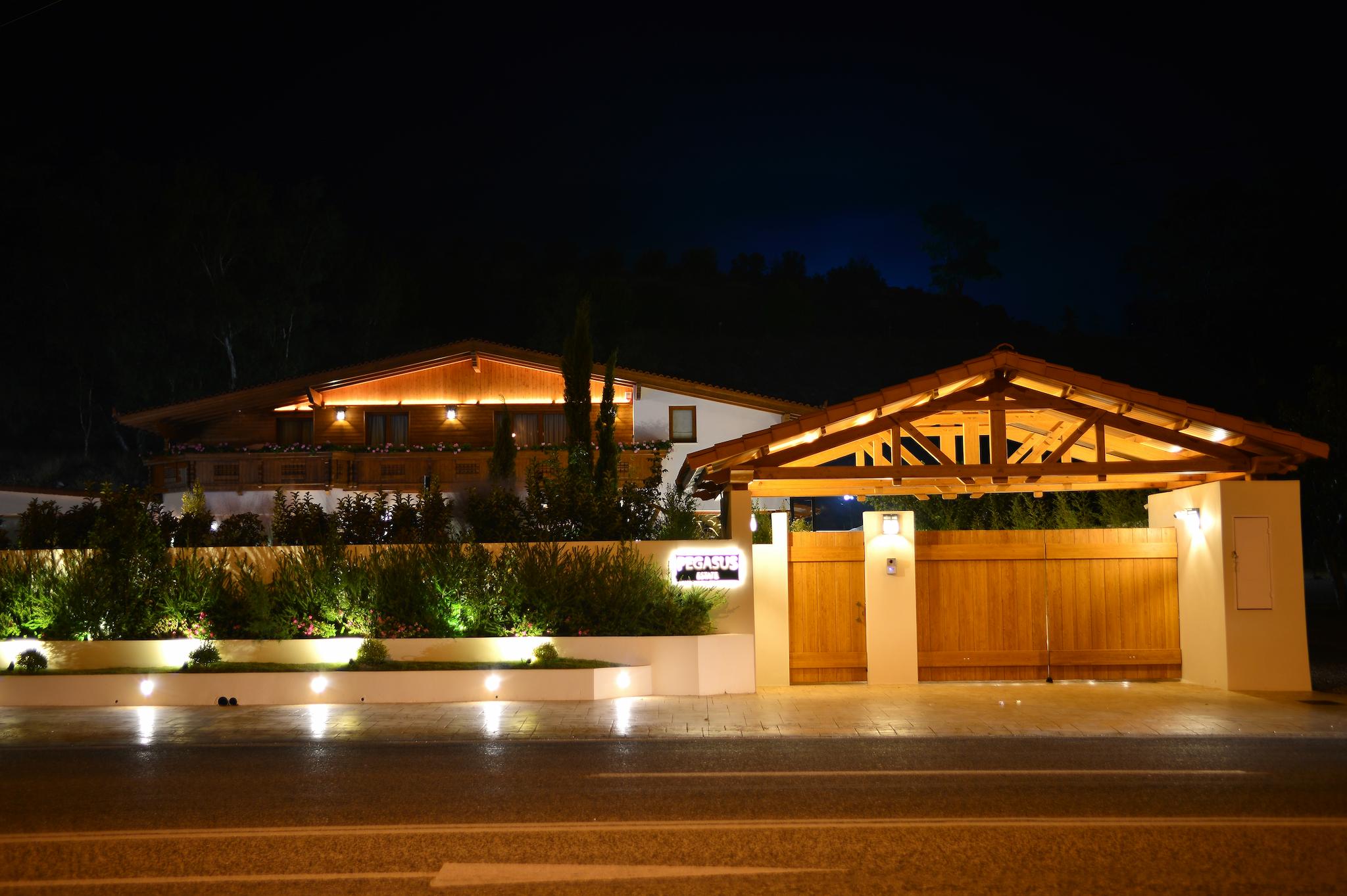 Εξωτερική φωτογραφία τραβηγμένη το βράδυ που φαίνονται τα φώτα. Είναι η πρόσοψη του σπιτιού με το στέγαστρο και την ξύλινη πόρτα και το ελβετικό σαλέ στο βάθος. Μπροστά βρίσκεται η παραλιακή λεωφόρο Αθηνών Σουνίου Στα μπροστινά παρτέρια που έχουμε φαίνεται η πρασινάδα όπως επίσης και τα φυτά