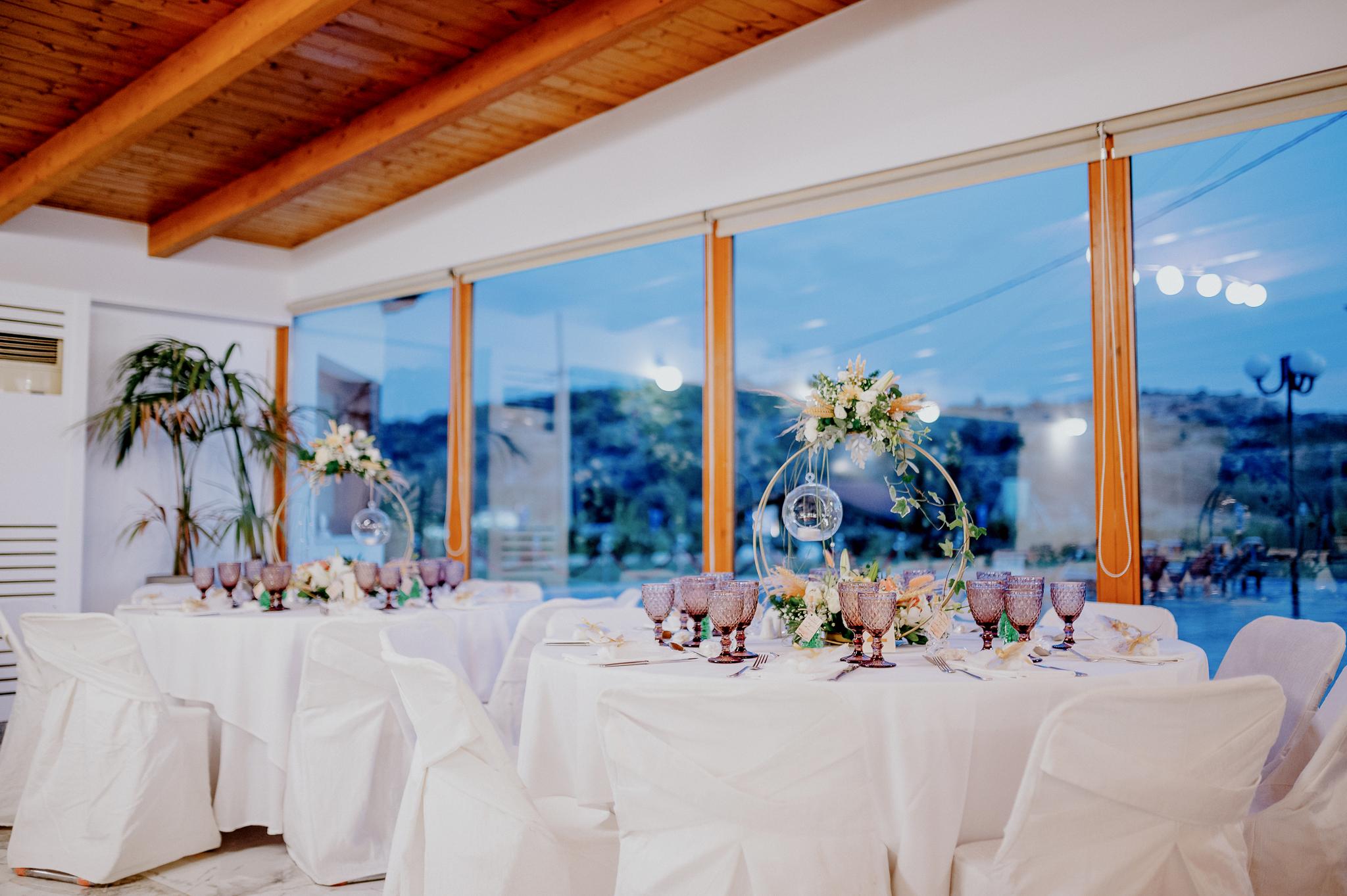 Απομακρυσμένη φωτογραφία στον εσωτερικό χώρο με ροτόντες και τραπέζια περιμετρικά από τις ροτόντες. Επάνω τα τραπέζια είναι στρωμένα με ποτήρια και μαχαιροπίρουνα. Στην μέση των τραπεζιών υπάρχει στολισμός όποιός αποτελείτε από γαρδένιας μαζί με λευκά και σομόν τριαντάφυλλα όπως και πρασινάδες. Στην μέση από το στρογγυλό που αποτελούνται κρέμεται ένα κεράκι . Στο βάθος φαίνονται οι τζαμαρίες του κτήματος που αφήνουν να δεις των έξω χώρο. Φαίνεται η ξύλινη στέγη στην οροφή που υπάρχει