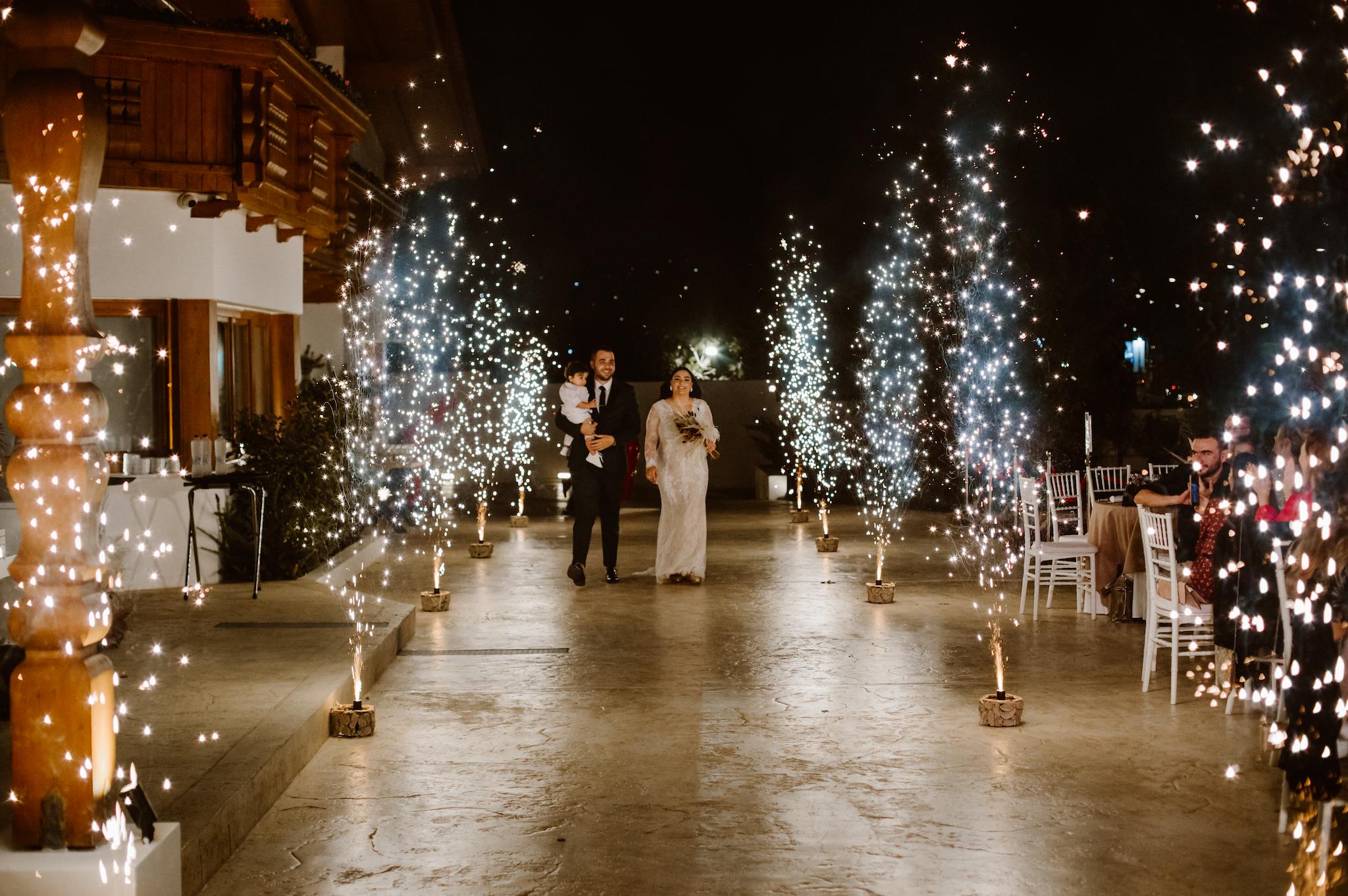 Ζευγάρι άνδρας που κρατάει ένα παιδί και γυναίκα με νυφικό το οποίο μπαίνει στο χώρο του κτήματος με επιδαπέδια βεγγαλικά το οποία είναι αναμμένα