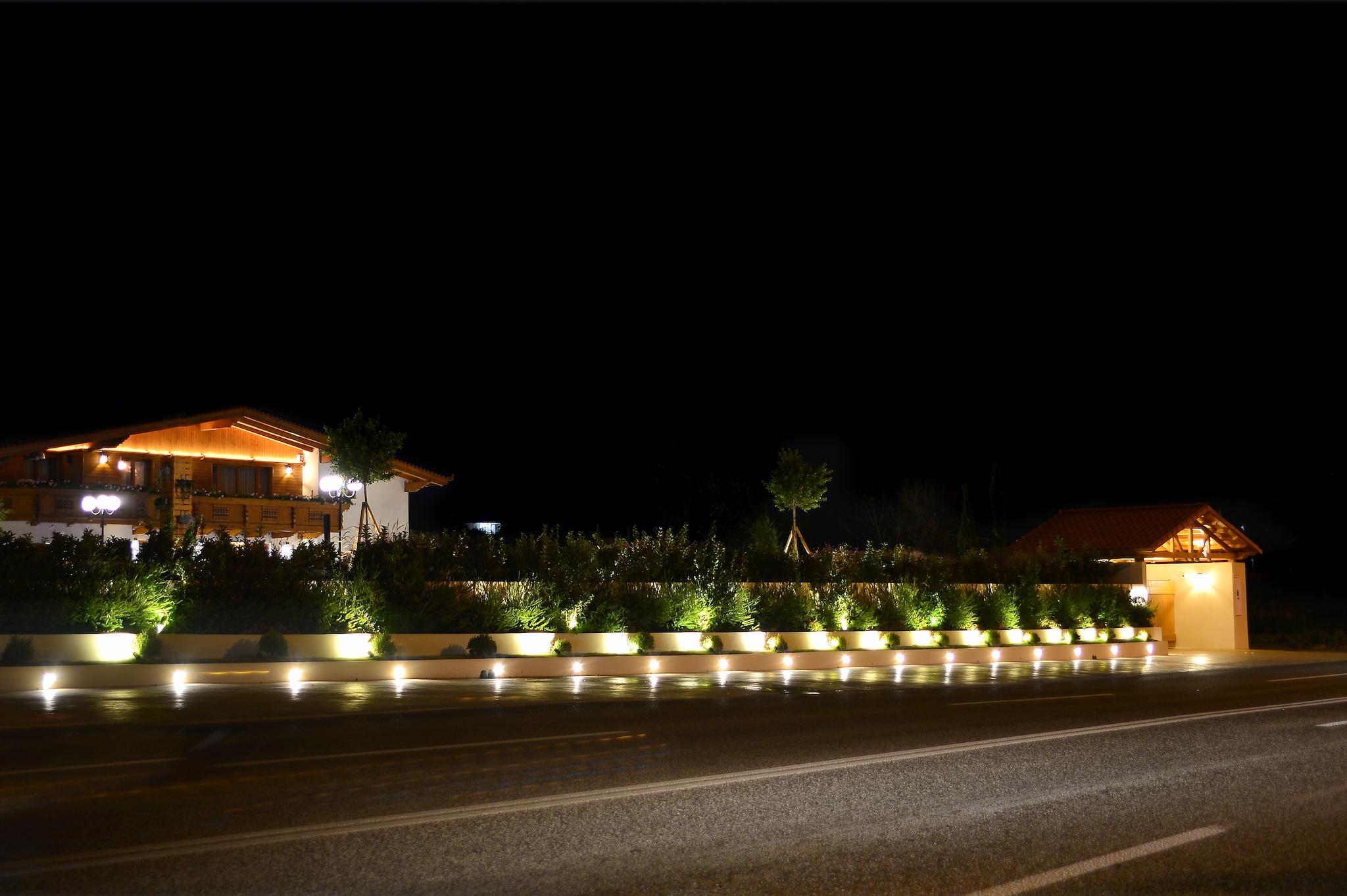 Εξωτερική φωτογραφία τραβηγμένη το βράδυ που φαίνονται τα φώτα. Είναι η πρόσοψη του σπιτιού με το στέγαστρο και την ξύλινη πόρτα και το ελβετικό σαλέ στο βάθος. Μπροστά βρίσκεται η παραλιακή λεωφόρο Αθηνών Σουνίου Στα μπροστινά παρτέρια που έχουμε φαίνεται η πρασινάδα όπως επίσης και τα φυτά. Η φωτογραφία είναι τραβηγμένη στην παραλιακή λεωφόρο Αθηνών Σουνίου