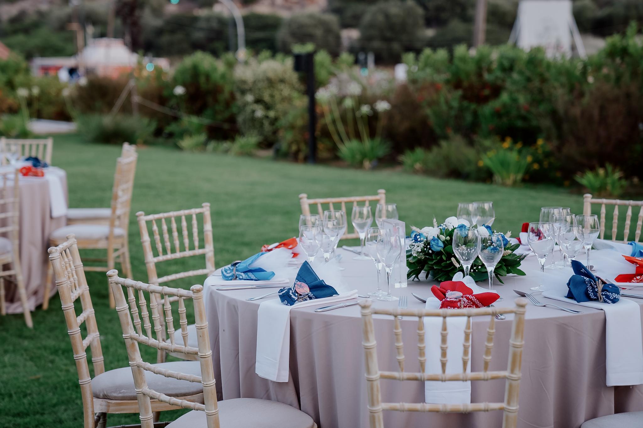 Κοντινή φωτογραφία στα τραπέζια του κτήματος , λουλούδια βρίσκονται επάνω στα τραπέζια τα οποία περιέχουν μπλε τριαντάφυλλα και λευκά όπως και γυψοφύλλι . Επάνω στο κάθε τραπέζι βρίσκονται τοποθετημένες και η μπομπονιέρες. Στο βάθος φαίνεται το γκαζόν όπως επίσης και και ο πράσινος κήπος με ταλουλούδια.