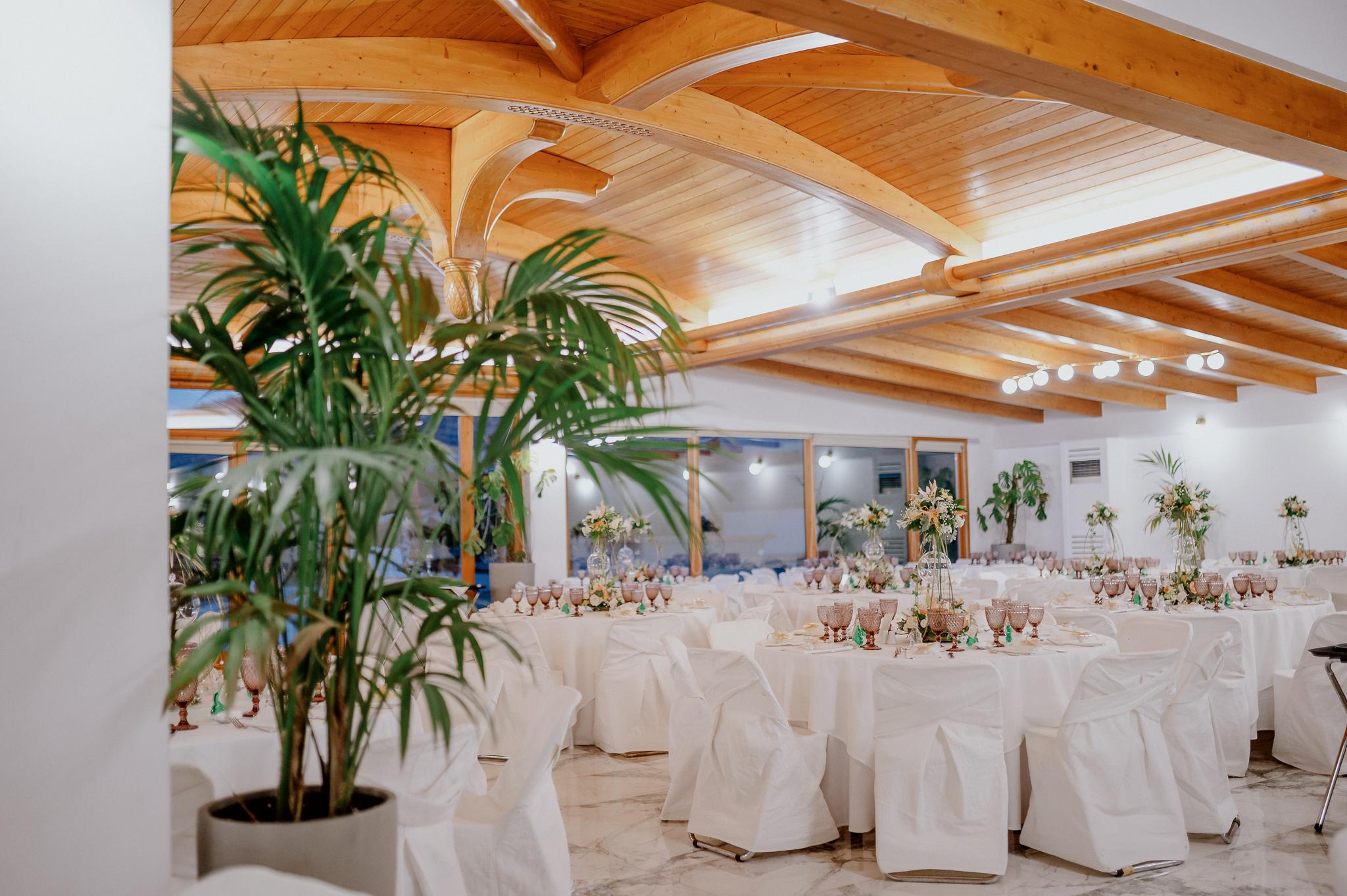 Απομακρυσμένη φωτογραφία στον εσωτερικό χώρο με ροτόντες και τραπέζια περιμετρικά από τις ροτόντες. Επάνω τα τραπέζια είναι στρωμένα με ποτήρια και μαχαιροπίρουνα. Στην μέση των τραπεζιών υπάρχει στολισμός όποιός αποτελείτε από γαρδένιας μαζί με λευκά και σομόν τριαντάφυλλα όπως και πρασινάδες. Στην μέση από το στρογγυλό που αποτελούνται κρέμεται ένα κεράκι . Στο βάθος φαίνονται οι τζαμαρίες του κτήματος που αφήνουν να δεις των έξω χώρο. Φαίνεται η ξύλινη στέγη στην οροφή που υπάρχει