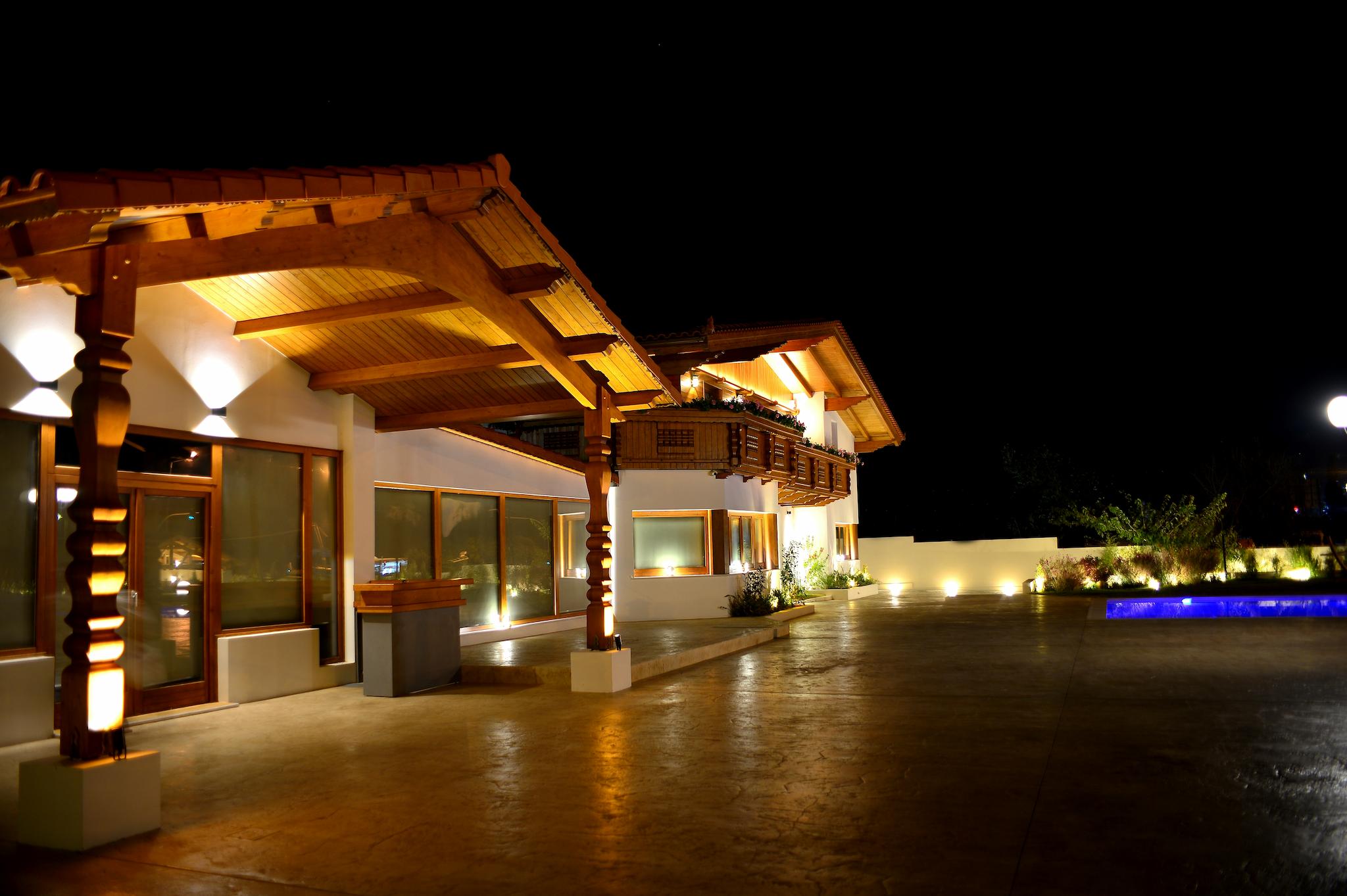 Φωτογραφία νυχτερινή η οποία δείχνει το ξύλινο ελβετικό σαλέ φωτισμένο. Στο βάθος φαίνεται και η πισίνα όπως επίσης και τον χώρο που τοποθετούνται τα τραπέζια.