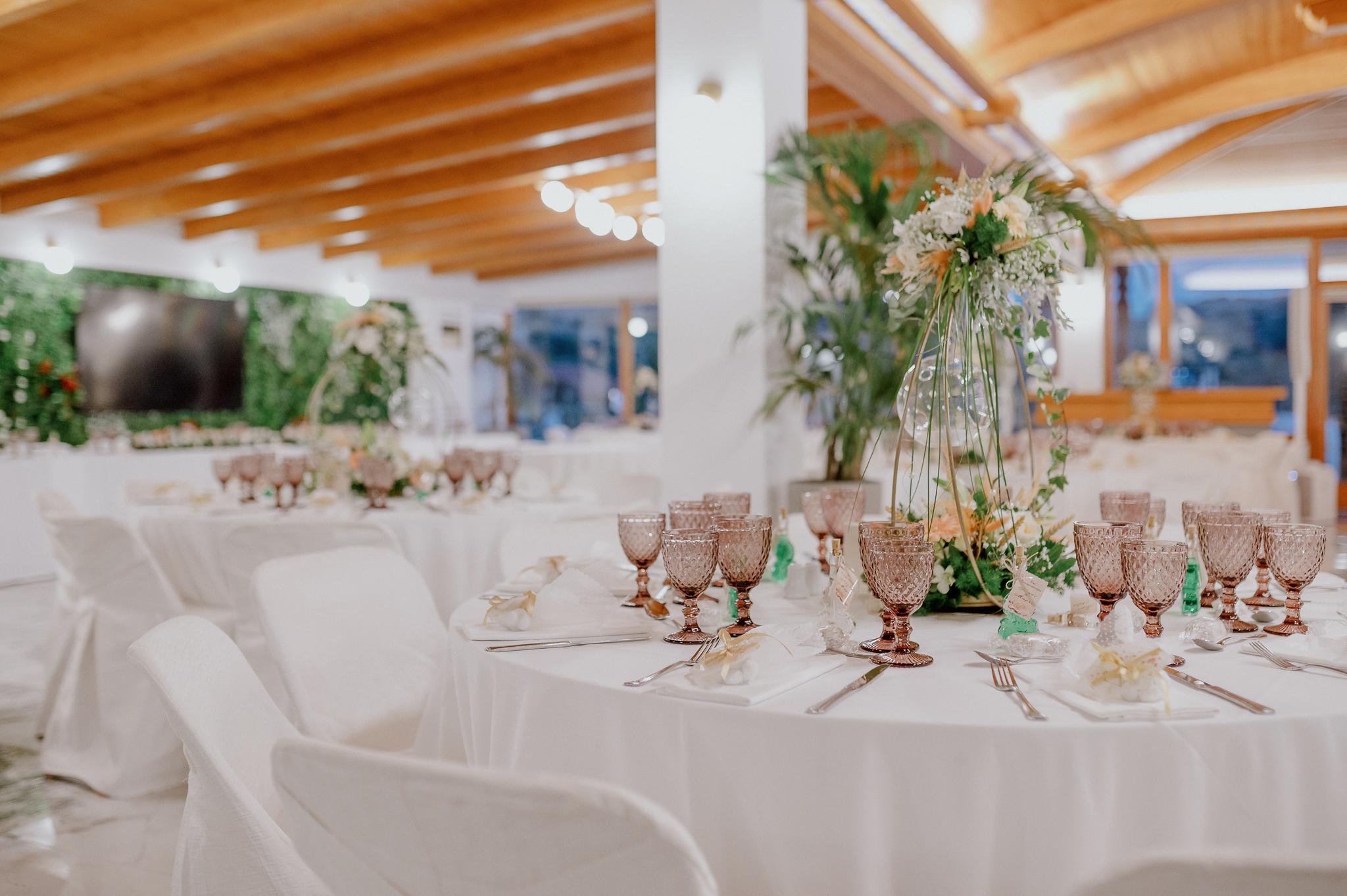 Απομακρυσμένη φωτογραφία στον εσωτερικό χώρο με ροτόντες και τραπέζια περιμετρικά από τις ροτόντες. Επάνω τα τραπέζια είναι στρωμένα από το catering με ποτήρια και μαχαιροπίρουνα. Στην μέση των τραπεζιών υπάρχει στολισμός όποιός αποτελείτε από γαρδένιας μαζί με λευκά και σομόν τριαντάφυλλα όπως και πρασινάδες. Στην μέση από το στρογγυλό που αποτελούνται κρέμεται ένα κεράκι . Στο βάθος φαίνονται οι τζαμαρίες του κτήματος που αφήνουν να δεις των έξω χώρο. Η οροφή της ξύλινης στέγης είναι ξύλινη