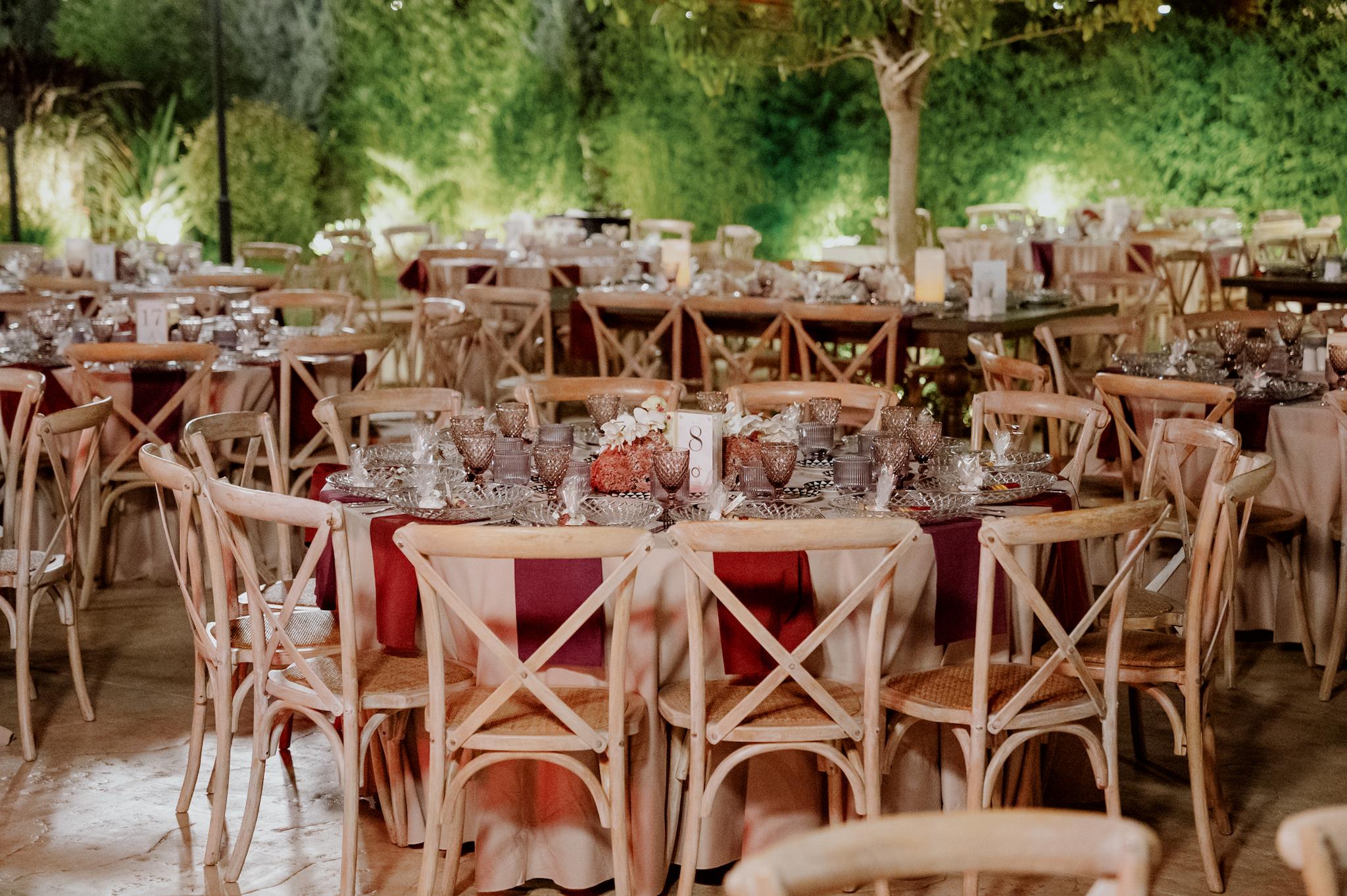 Κοντινή φωτογραφία με ροτόντες τραπέζια περιμετρικά από τα τραπέζια είναι ξύλινες καρέκλες. Τα τραπέζια έχουν μπεζ τραπεζομάντηλα και επάνω μπορντό ατομικές πετσέτες. Στο βάθος είναι το αυτοκινούμενο βαν μπαρ το οποίο το χρησιμοποιούν οι καλεσμένοι για να παίρνουν ποτό . Στο βάθος φαίνεται το γκαζόν αλλά και τα φυτά που έχει το κτήμα. Στο βάθος φαίνεται το πράσινο και ο κήπος