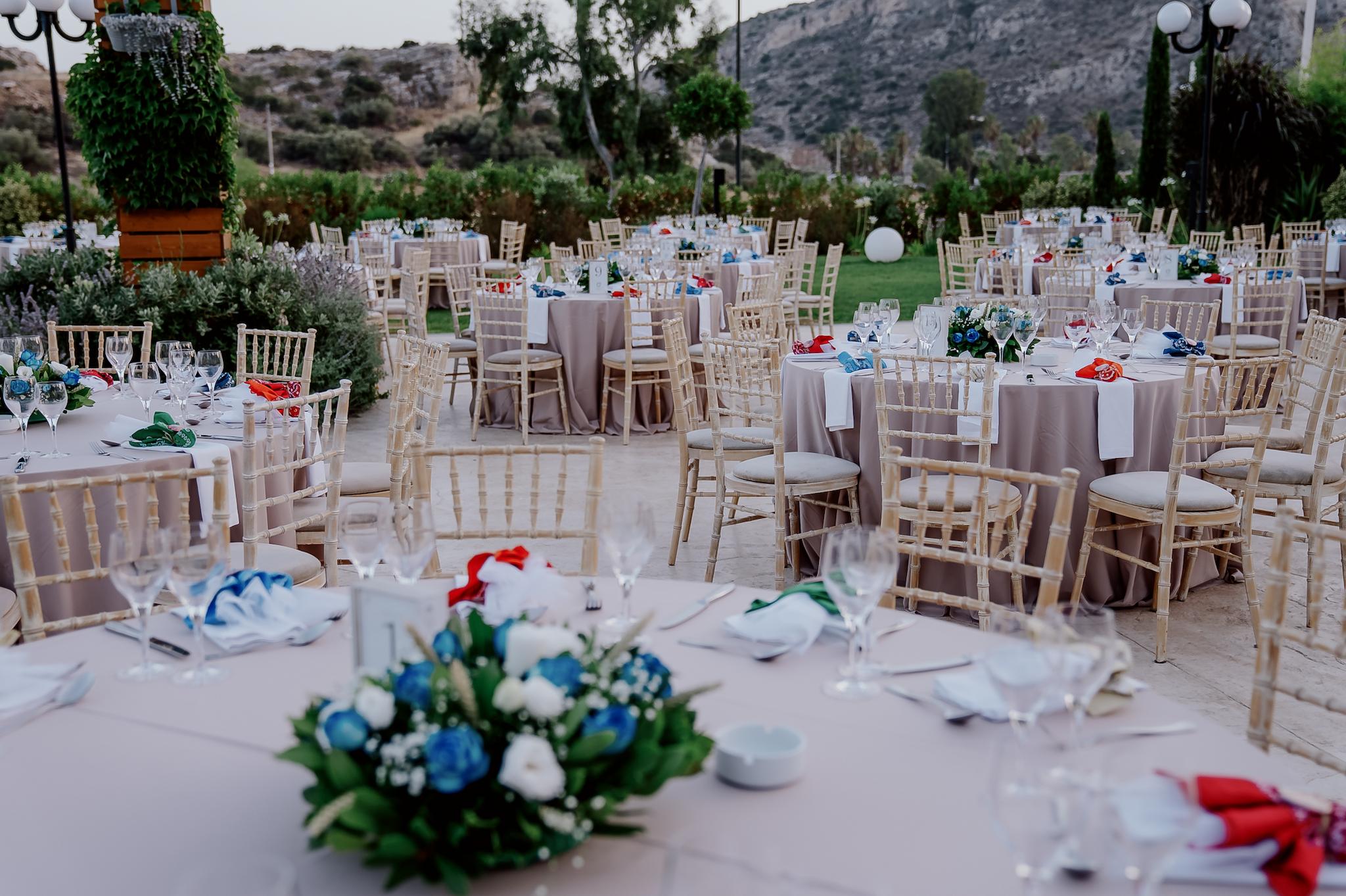 Μακρινή φωτογραφία στα τραπέζια του κτήματος , λουλούδια βρίσκονται επάνω στα τραπέζια τα οποία περιέχουν μπλε τριαντάφυλλα και λευκά . Επάνω στο κάθε τραπέζι βρίσκονται τοποθετημένες και η μπομπονιέρες. Στο βάθος φαίνεται ο πράσινος κήπος του κτήματος