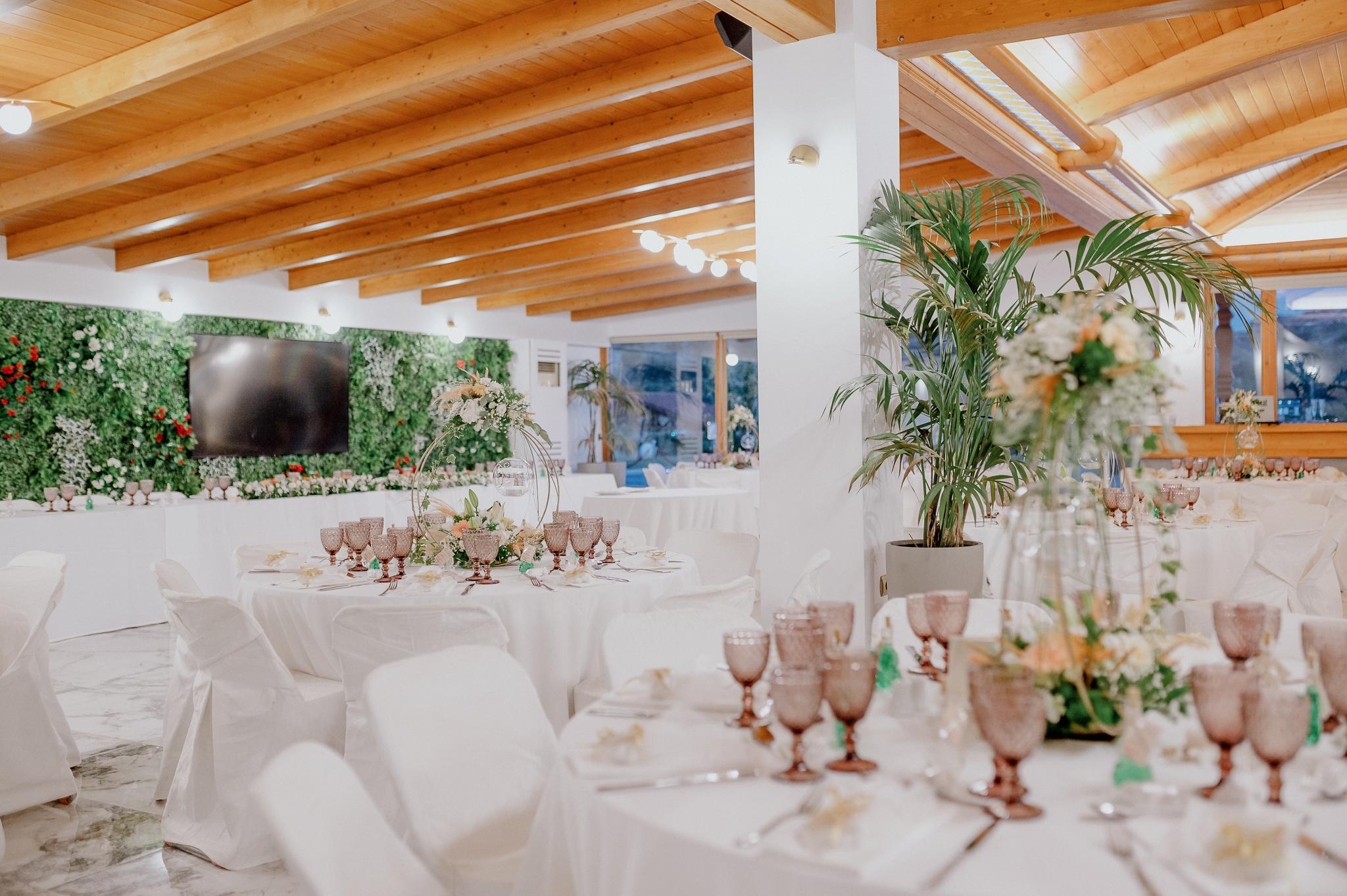 Απομακρυσμένη φωτογραφία στον εσωτερικό χώρο με ροτόντες και τραπέζια περιμετρικά από τις ροτόντες. Επάνω τα τραπέζια είναι στρωμένα με ποτήρια και μαχαιροπίρουνα. Στην μέση των τραπεζιών υπάρχει στολισμός όποιός αποτελείτε από γαρδένιας μαζί με λευκά και σομόν τριαντάφυλλα όπως και πρασινάδες. Στην μέση από το στρογγυλό που αποτελούνται κρέμεται ένα κεράκι . Στο βάθος φαίνονται οι τζαμαρίες του κτήματος που αφήνουν να δεις των έξω χώρο