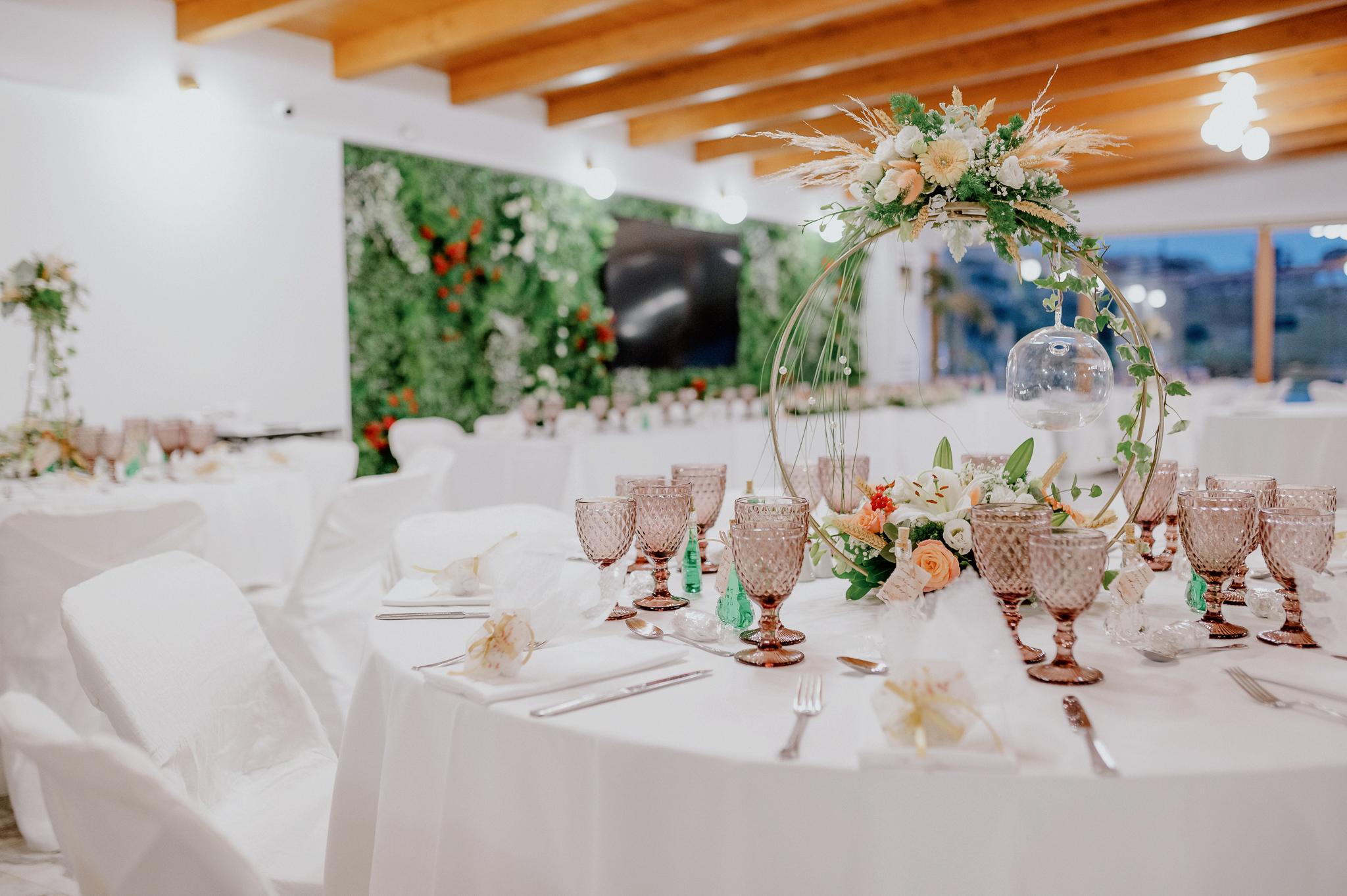 Απομακρυσμένη φωτογραφία στον εσωτερικό χώρο με ροτόντες και τραπέζια περιμετρικά από τις ροτόντες. Επάνω τα τραπέζια είναι στρωμένα από το catering με ποτήρια και μαχαιροπίρουνα. Στην μέση των τραπεζιών υπάρχει στολισμός όποιός αποτελείτε από γαρδένιας μαζί με λευκά και σομόν τριαντάφυλλα όπως και πρασινάδες. Στην μέση από το στρογγυλό που αποτελούνται κρέμεται ένα κεράκι . Στο βάθος φαίνονται οι τζαμαρίες του κτήματος που αφήνουν να δεις των έξω χώρο. Η οροφή της ξύλινης στέγης είναι ξύλινη