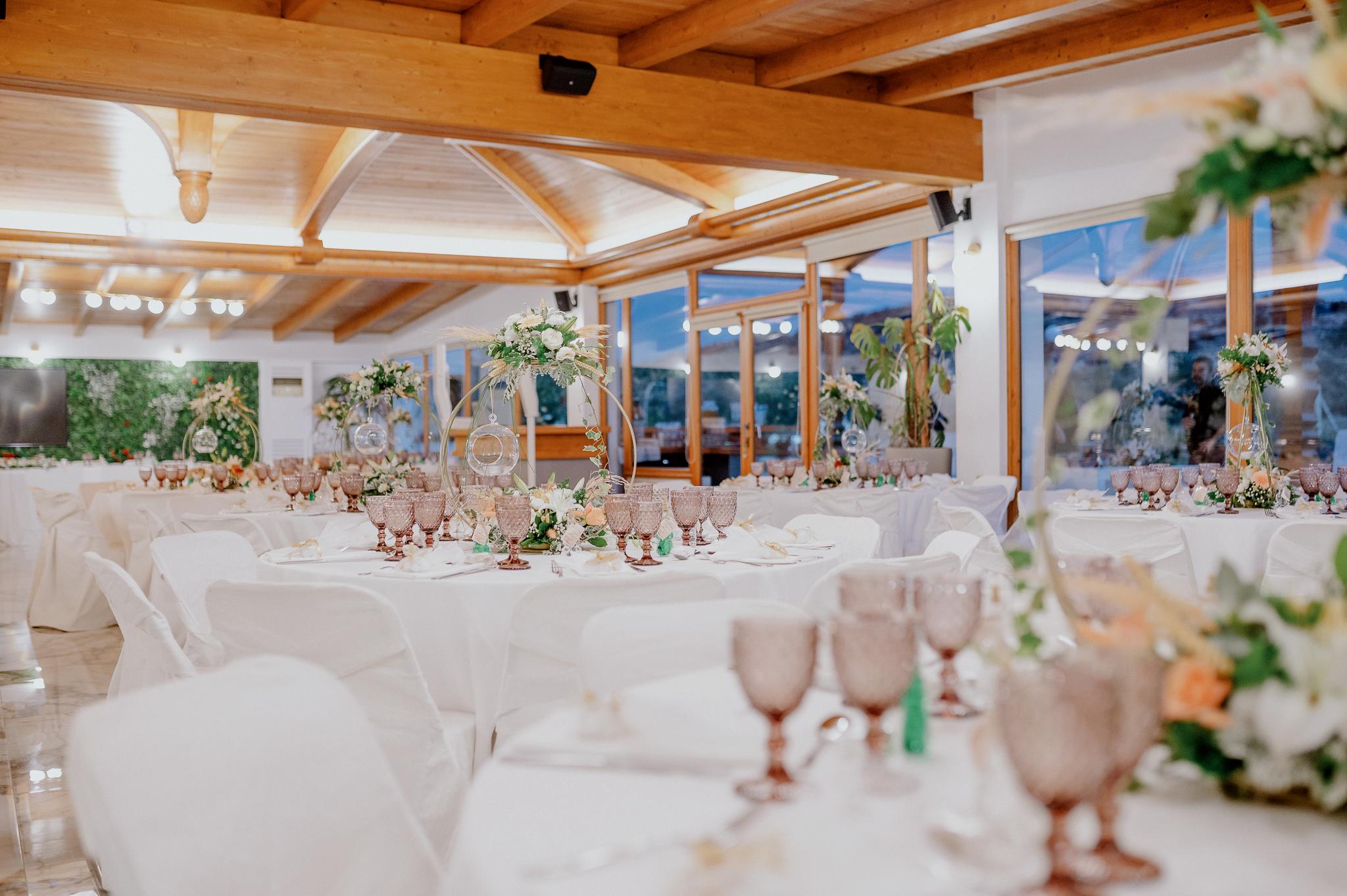 Απομακρυσμένη φωτογραφία στον εσωτερικό χώρο με ροτόντες και τραπέζια περιμετρικά από τις ροτόντες. Επάνω τα τραπέζια είναι στρωμένα με ποτήρια και μαχαιροπίρουνα. Στην μέση των τραπεζιών υπάρχει στολισμός όποιός αποτελείτε από γαρδένιας μαζί με λευκά και σομόν τριαντάφυλλα όπως και πρασινάδες. Στην μέση από το στρογγυλό που αποτελούνται κρέμεται ένα κεράκι . Στο βάθος φαίνονται οι τζαμαρίες του κτήματος που αφήνουν να δεις των έξω χώρο