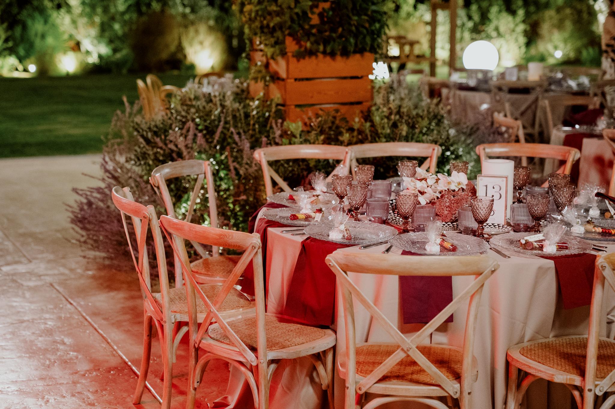 Κοντινή φωτογραφία με ροτόντες τραπέζια περιμετρικά από τα τραπέζια είναι ξύλινες καρέκλες. Τα τραπέζια έχουν μπεζ τραπεζομάντηλα και επάνω μπορντό ατομικές πετσέτες. Στο βάθος είναι το αυτοκινούμενο βαν μπαρ το οποίο το χρησιμοποιούν οι καλεσμένοι για να παίρνουν ποτό . Στο βάθος φαίνεται το γκαζόν αλλά και τα φυτά που έχει το κτήμα.