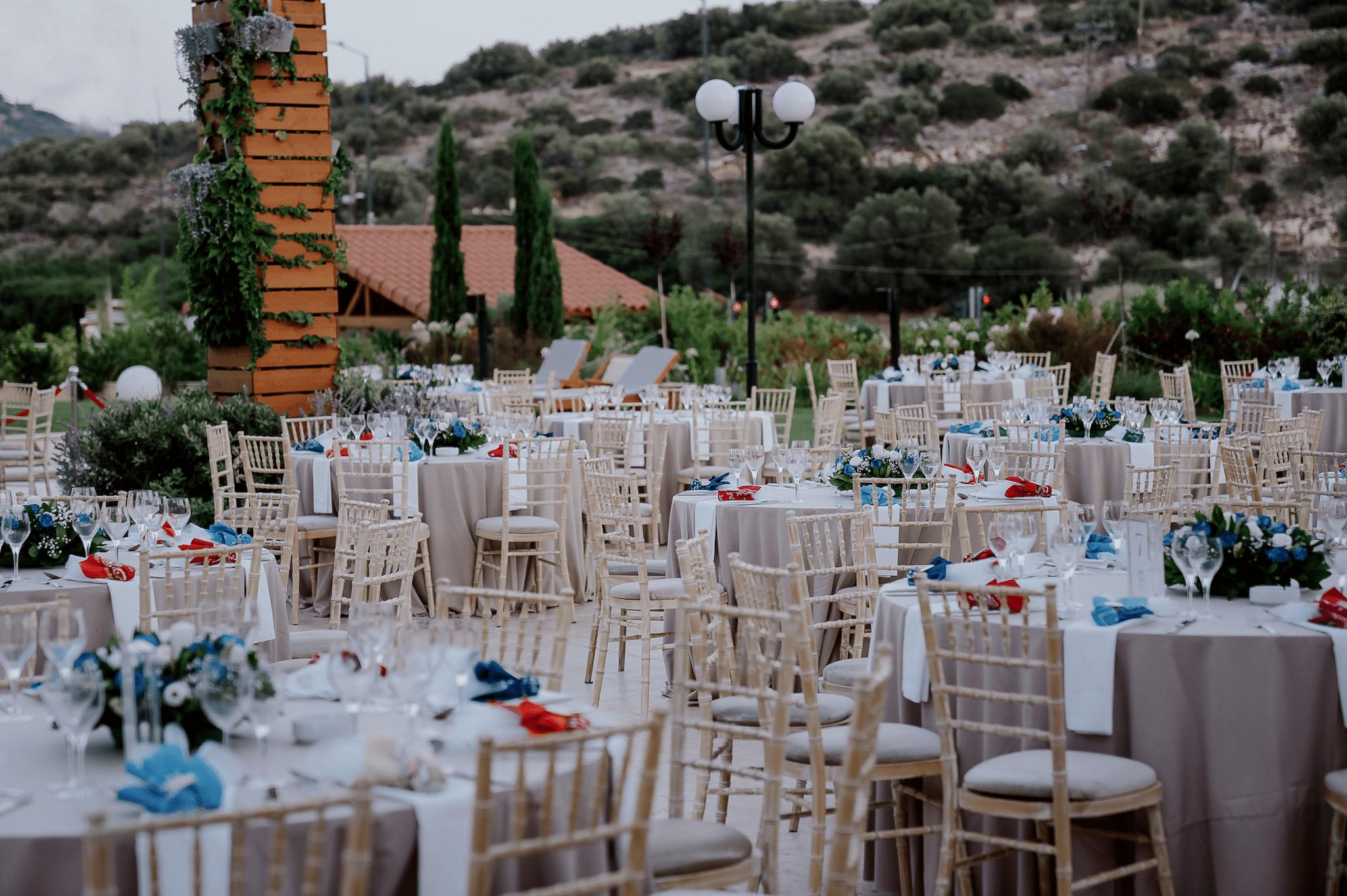 Μακρινό πλάνο με φόντο το πράσινο γκαζόν με φόντο τα φυτά. Υπάρχουν στρωμένες ροτόντες όπου περιμετρικά έχουν tifanny καρέκλες. Στο κέντρο του κάθε τραπεζιού υπάρχει στολισμός ο οποίος αποτελείτε από μπλε και λευκά τριαντάφυλλα και μια μπομπονιέρα ανά άτομο