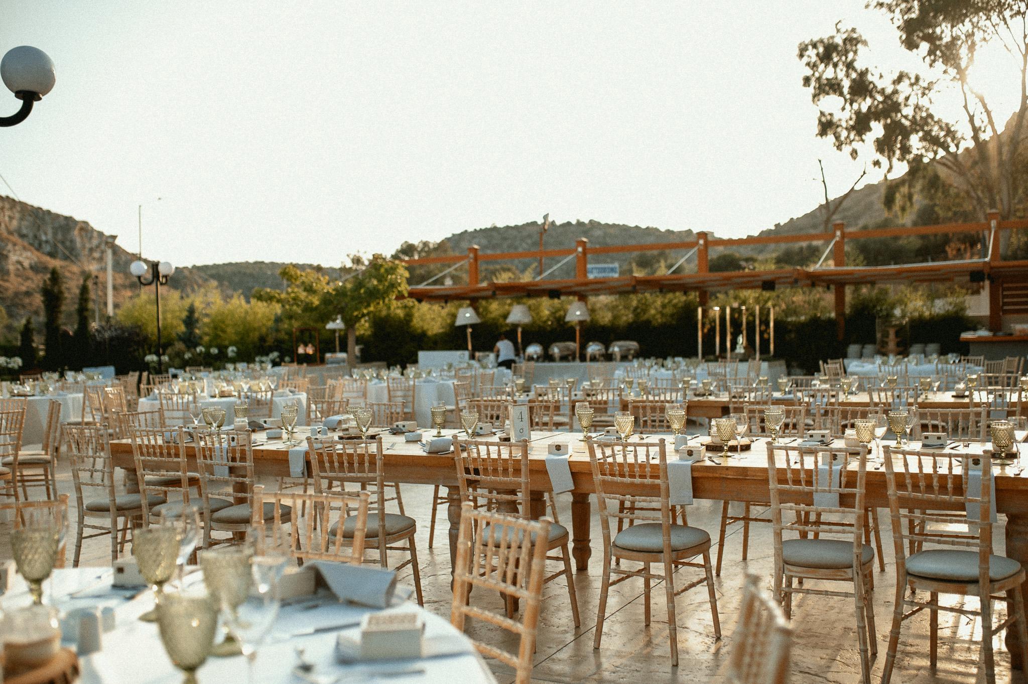 Απομακρυσμένη φωτογραφία πρωινή με στρωμένες ροτόντες και μοναστηριακά τραπέζια. Περιμετρικά από τα τραπέζια είναι στημένες οι καρέκλες ξύλινες tiffany τις οποίες τις έχει στήσει το catering . Στο βάθος τις φωτογραφίας φαίνεται η ξύλινη πέργκολα που από κάτω έχει στήσει το cateirng