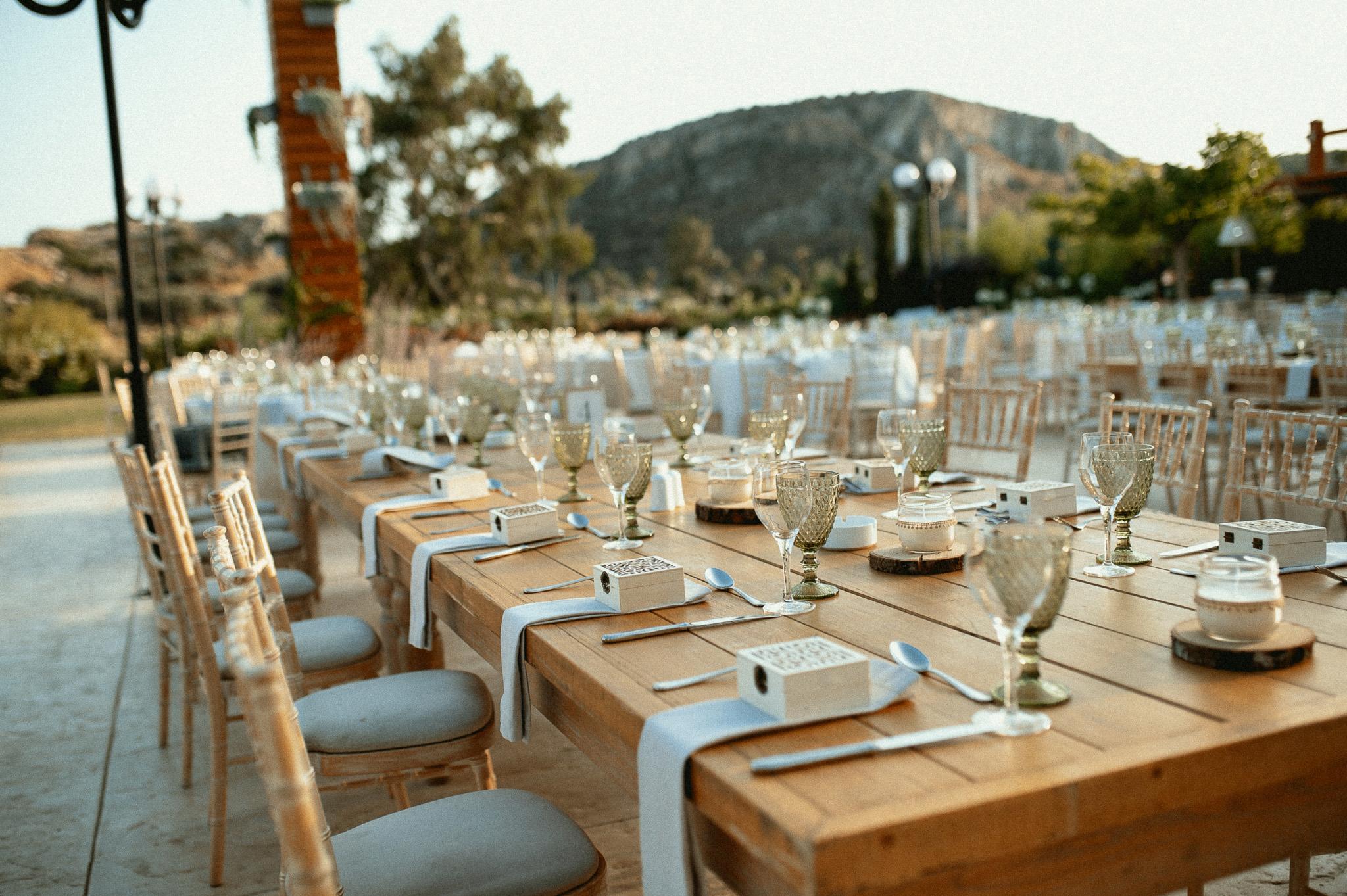 Μοναστηριακό τραπέζι με πράσινα ποτήρια και λευκές πετσέτες ατομικές με τις μπομπονιέρες επάνω στις πετσέτες και καρέκλες ξύλινες. Επάνω στα τραπέζια το catering έχει στρώση με ποτήρια και μαχαιροπίρουνα όπως επίσης υπάρχει στο κάθε άτομα και μια μπομπονιέρα . Στην μέση του τραπεζιού υπάρχει ένα ξύλινος κορμός που επάνω που έχει κεράκι και υπάρχει και ένα βάζο το οποίο μέσα έχει pampas σε χρώματα μπεζ και ένα τριαντάφυλλο λευκό