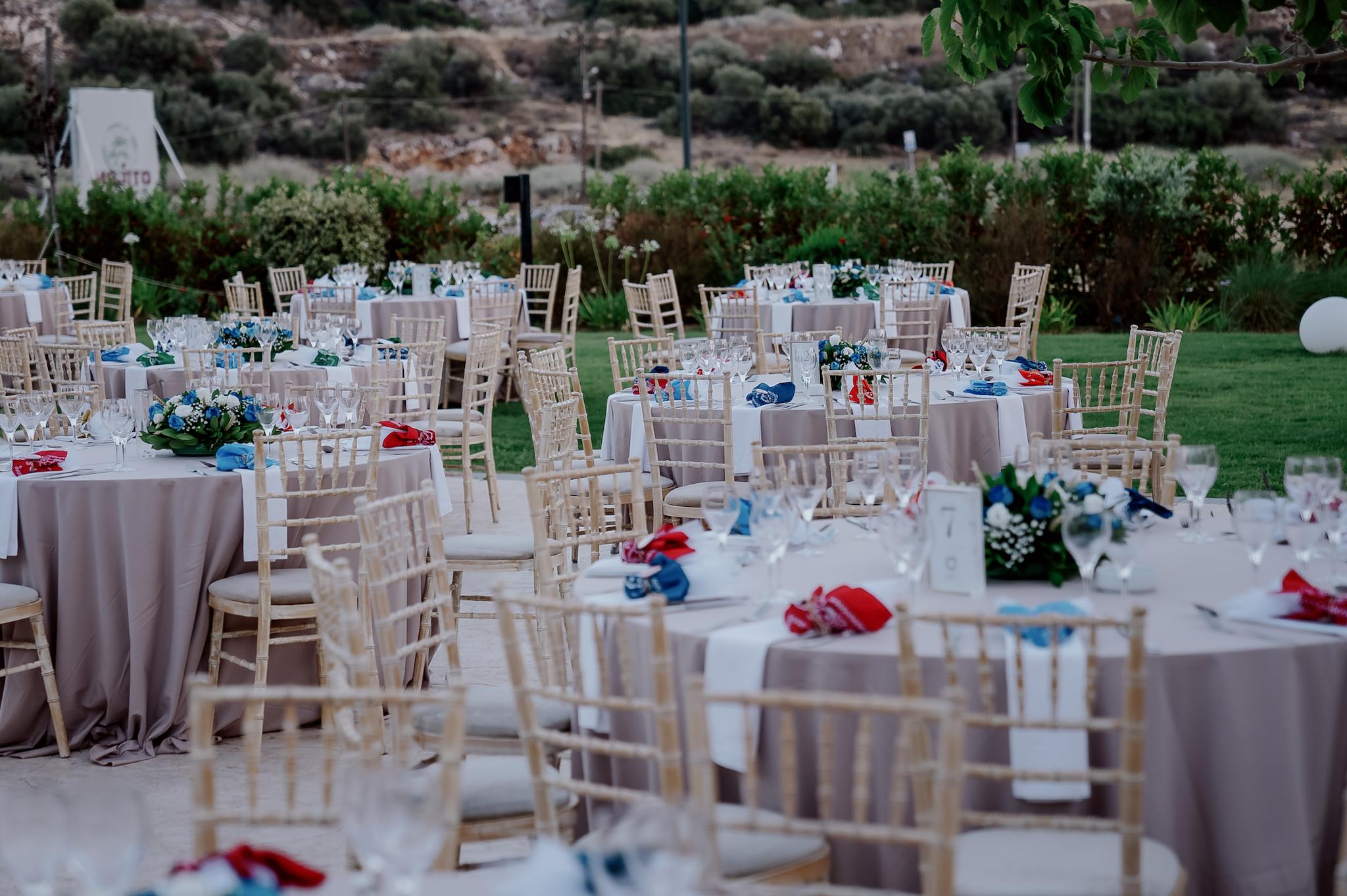 Μακρινό πλάνο με φόντο το πράσινο γκαζόν με φόντο τα φυτά. Υπάρχουν στρωμένες ροτόντες όπου περιμετρικά έχουν tifanny καρέκλες. Στο κέντρο του κάθε τραπεζιού υπάρχει στολισμός ο οποίος αποτελείτε από μπλε και λευκά τριαντάφυλλα και μια μπομπονιέρα ανά άτομο