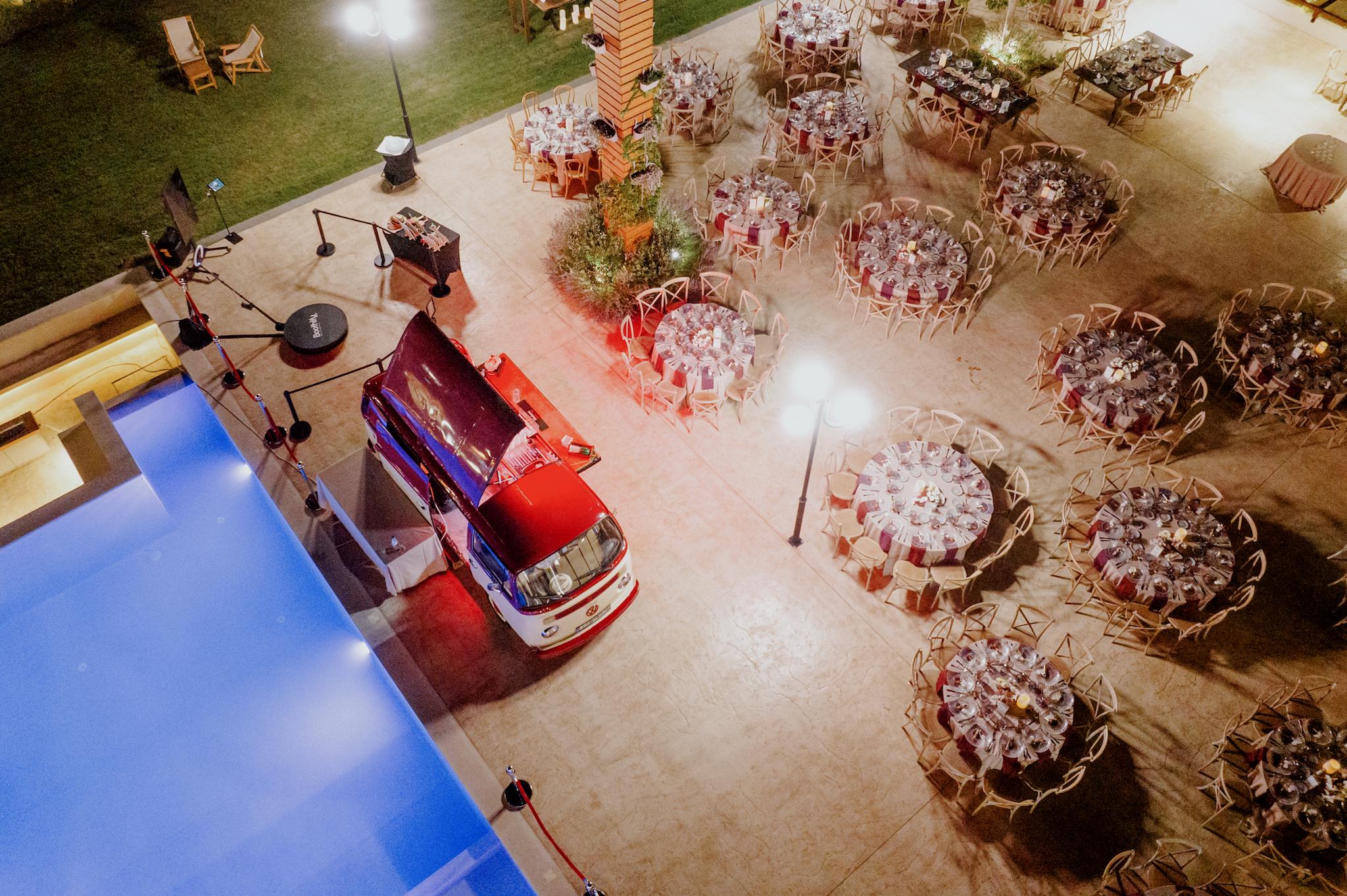 Φωτογραφία από ψηλά που φαίνεται το βαν μπαρ και τα τραπέζια. Το βαν βρίσκεται δίπλα στην πισίνα. Μπροστά του υπάρχουν τα τραπέζια ροτόντες αλλά και τα μοναστηριακά. στρωμένα έτοιμα με καρέκλες και μαχαιροπίρουνα πιάτα και τραπεζομάντηλα