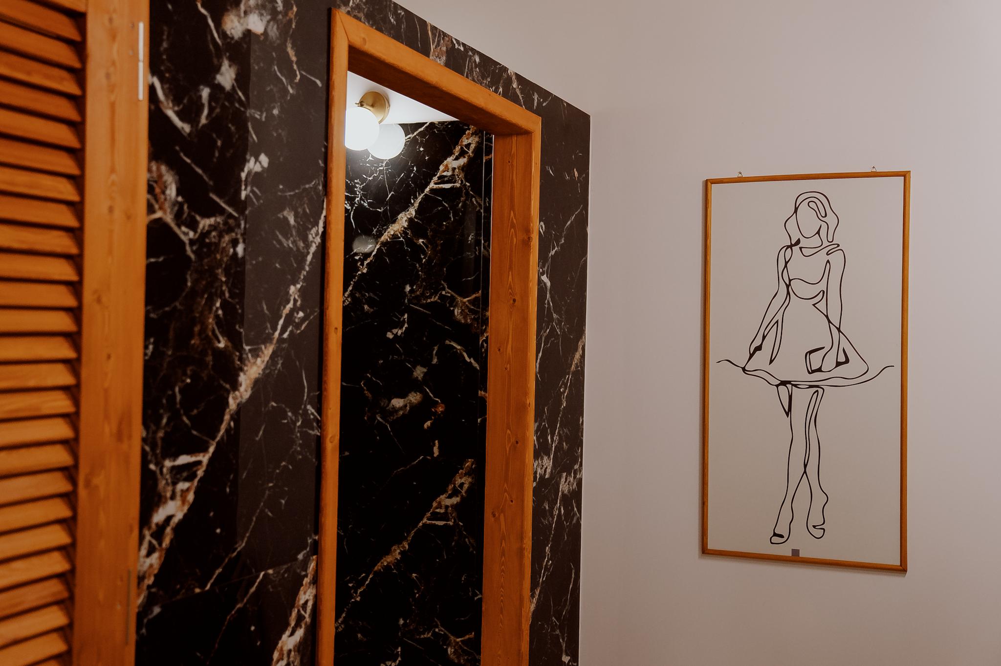 απεικονίζει ένα κάδρο με μία γυναίκα το οποίο είναι η ένδειξη για τις τουαλέτες. Φαίνονται τα γρανιτοπλακάκια τα οποία είναι λεύκα και μαύρα και ξύλινες πόρτες