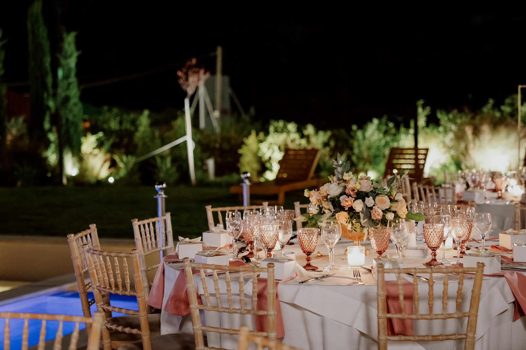 Στολισμός στα τραπέζια με τριαντάφυλλα σάπιο μήλο και λευκά με πολύχρωμα ποτήρια σε ροζ αποχρώσεις και καρέκλες tiffany στο βάθος φαίνεται το πολύχρωμα τοπίο από πράσινο και τα φυτά