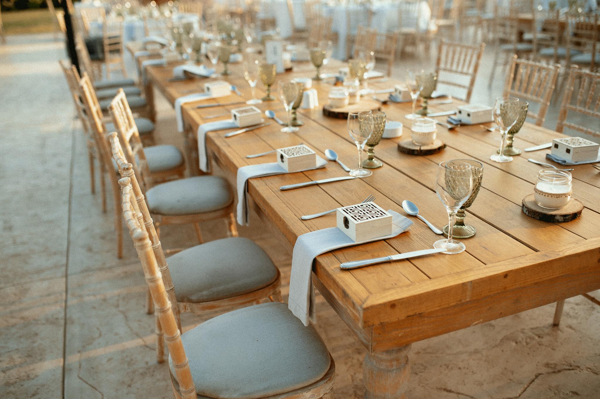 Μοναστηριακό τραπέζι με πράσινα ποτήρια και λευκές πετσέτες ατομικές με τις μπομπονιέρες επάνω στις πετσέτες και καρέκλες ξύλινες. Επάνω στα τραπέζια το catering έχει στρώση με ποτήρια και μαχαιροπίρουνα όπως επίσης υπάρχει στο κάθε άτομα και μια μπομπονιέρα . Στην μέση του τραπεζιού υπάρχει ένα ξύλινος κορμός που επάνω που έχει κεράκι και υπάρχει και ένα βάζο το οποίο μέσα έχει pampas σε χρώματα μπεζ και ένα τριαντάφυλλο λευκό