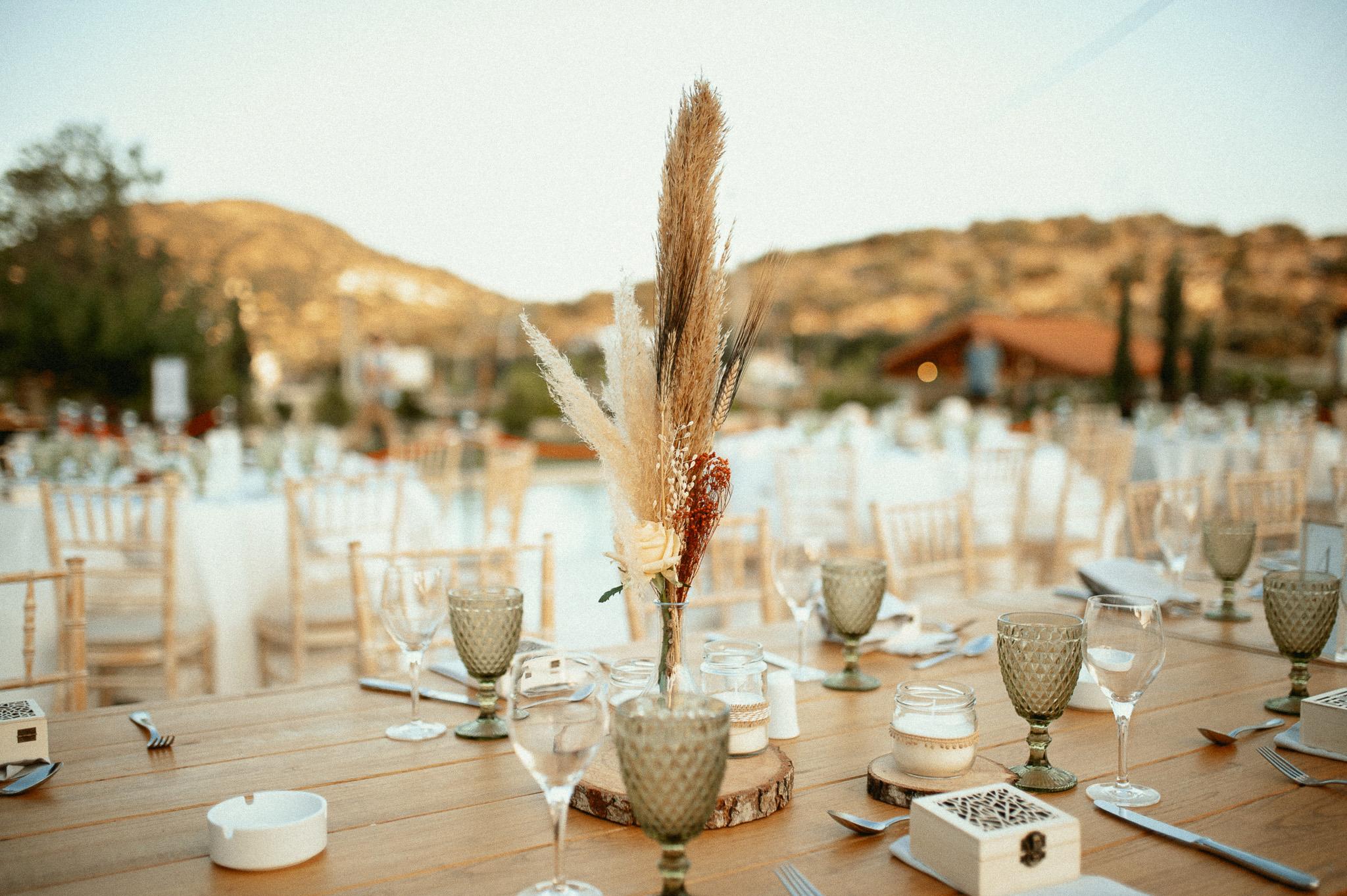 Μοναστηριακό τραπέζι με πράσινα ποτήρια και λευκές πετσέτες ατομικές με τις μπομπονιέρες επάνω στις πετσέτες και καρέκλες ξύλινες. Επάνω στα τραπέζια το catering έχει στρώση με ποτήρια και μαχαιροπίρουνα όπως επίσης υπάρχει στο κάθε άτομα και μια μπομπονιέρα . Στην μέση του τραπεζιού υπάρχει ένα ξύλινος κορμός που επάνω που έχει κεράκι και υπάρχει και ένα βάζο το οποίο μέσα έχει pampas σε χρώματα μπεζ και ένα τριαντάφυλλο λευκό