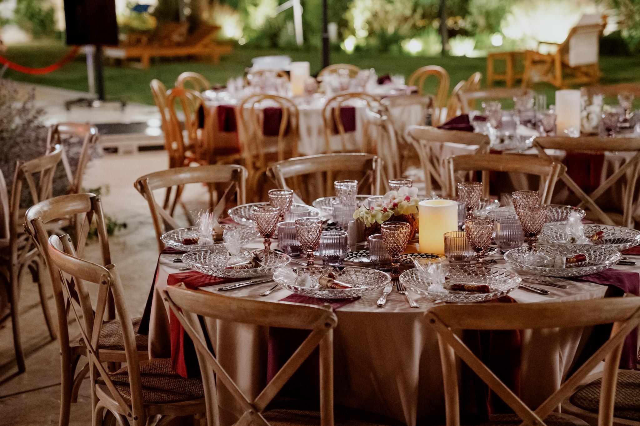 Κοντινή φωτογραφία με ροτόντες τραπέζια περιμετρικά από τα τραπέζια είναι ξύλινες καρέκλες. Τα τραπέζια έχουν μπεζ τραπεζομάντηλα και επάνω μπορντό ατομικές πετσέτες. Υπάρχουν οι μπομπονιέρες επάνω στο κάθε άτομο όπως επίσης και ένα κερί στην μέση του κάθε τραπεζιού.