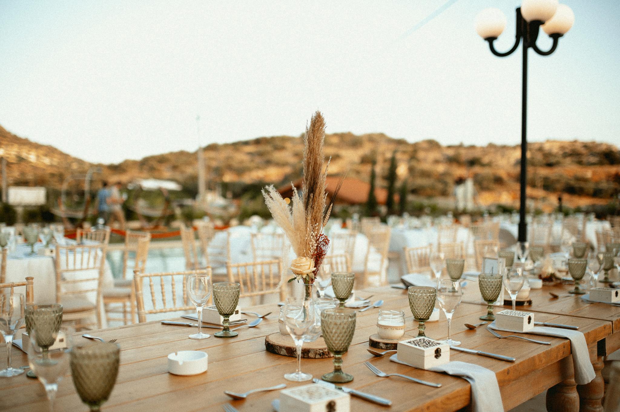 Μοναστηριακό τραπέζι με πράσινα ποτήρια και λευκές πετσέτες ατομικές με τις μπομπονιέρες επάνω στις πετσέτες και καρέκλες ξύλινες. Επάνω στα τραπέζια το catering έχει στρώση με ποτήρια και μαχαιροπίρουνα όπως επίσης υπάρχει στο κάθε άτομα και μια μπομπονιέρα . Στην μέση του τραπεζιού υπάρχει ένα ξύλινος κορμός που επάνω που έχει κεράκι και υπάρχει και ένα βάζο το οποίο μέσα έχει pampas σε χρώματα μπεζ και ένα τριαντάφυλλο λευκό