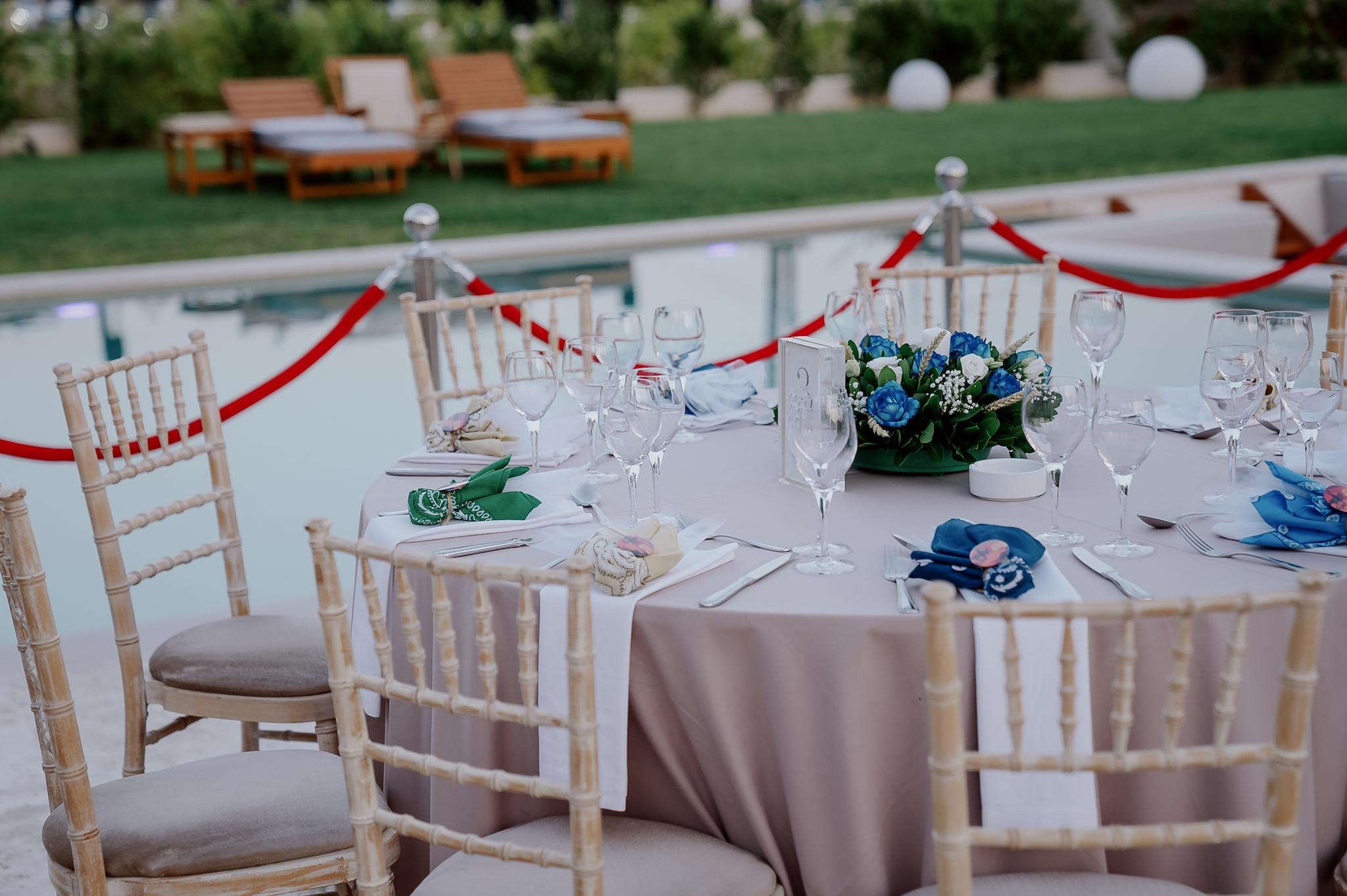 Μακρινή φωτογραφία στα τραπέζια του κτήματος , Λουλούδια βρίσκονται επάνω στα τραπέζια τα οποία περιέχουν μπλε τριαντάφυλλα και λευκά . Επάνω στο κάθε τραπέζι βρίσκονται τοποθετημένες και η μπομπονιέρες έχει στήσει το catering. Περιμετρικά από τις ροτόντες βρίσκονται οι tiffany ξύλινες καρέκλες