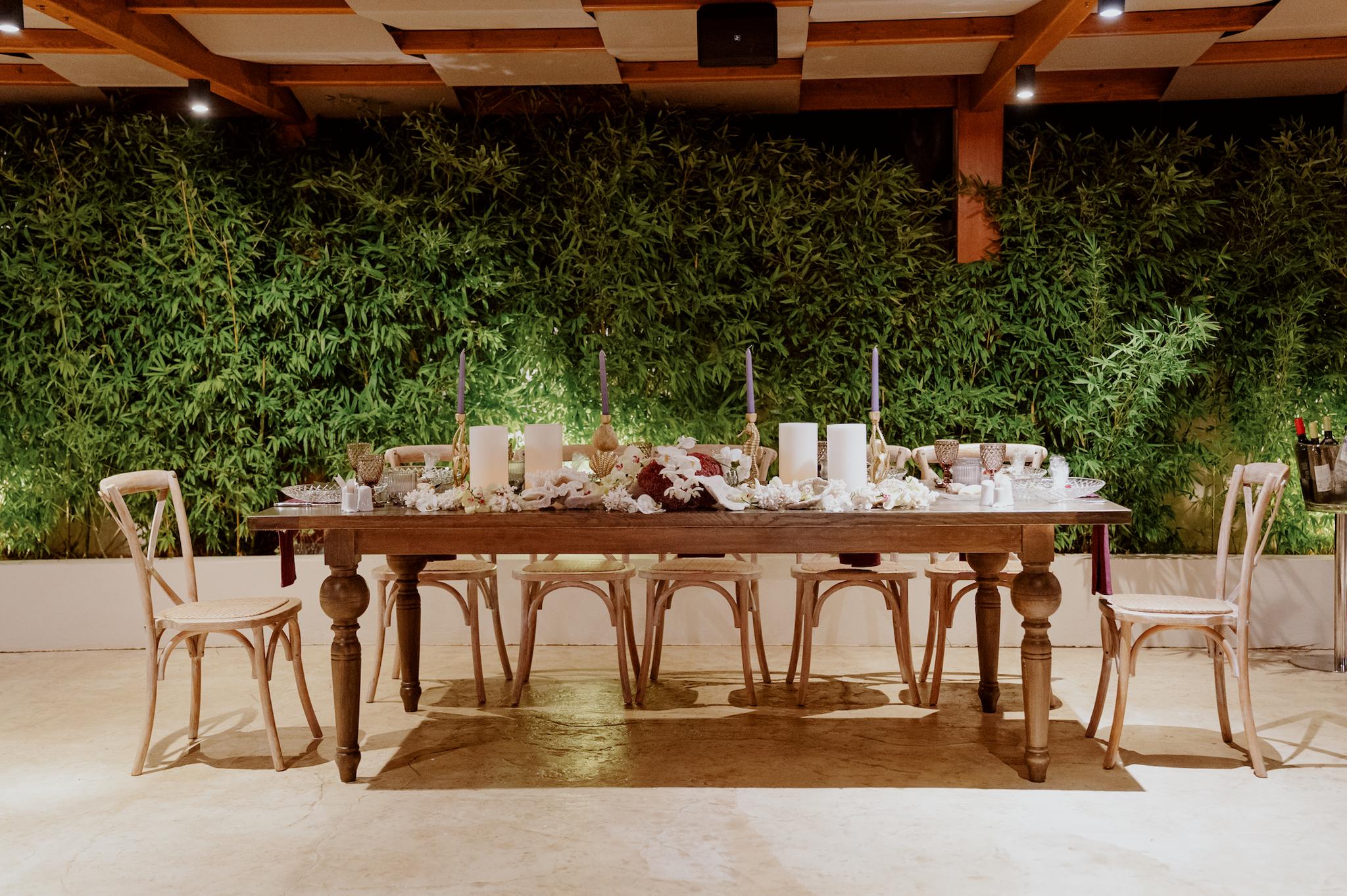 Μοναστηριακό τραπέζι κάτω από την πέργκολα περιμετρικά έχει καρέκλες ξύλινες τις οποίες τις έχει φέρει το catering επάνω στο τραπέζι έχει στολισμό με βότσαλα μπορντό και μπορντό ατομικές πετσέτες και διάφορα κεράκια . Στο φόντο έχει πράσινο τοπία απο φυτά