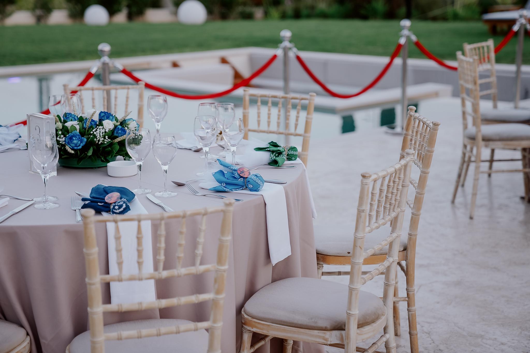 Μακρινή φωτογραφία στα τραπέζια του κτήματος , Λουλούδια βρίσκονται επάνω στα τραπέζια τα οποία περιέχουν μπλε τριαντάφυλλα και λευκά . Επάνω στο κάθε τραπέζι βρίσκονται τοποθετημένες και η μπομπονιέρες έχει στήσει το catering. Περιμετρικά από τις ροτόντες βρίσκονται οι tiffany ξύλινες καρέκλες