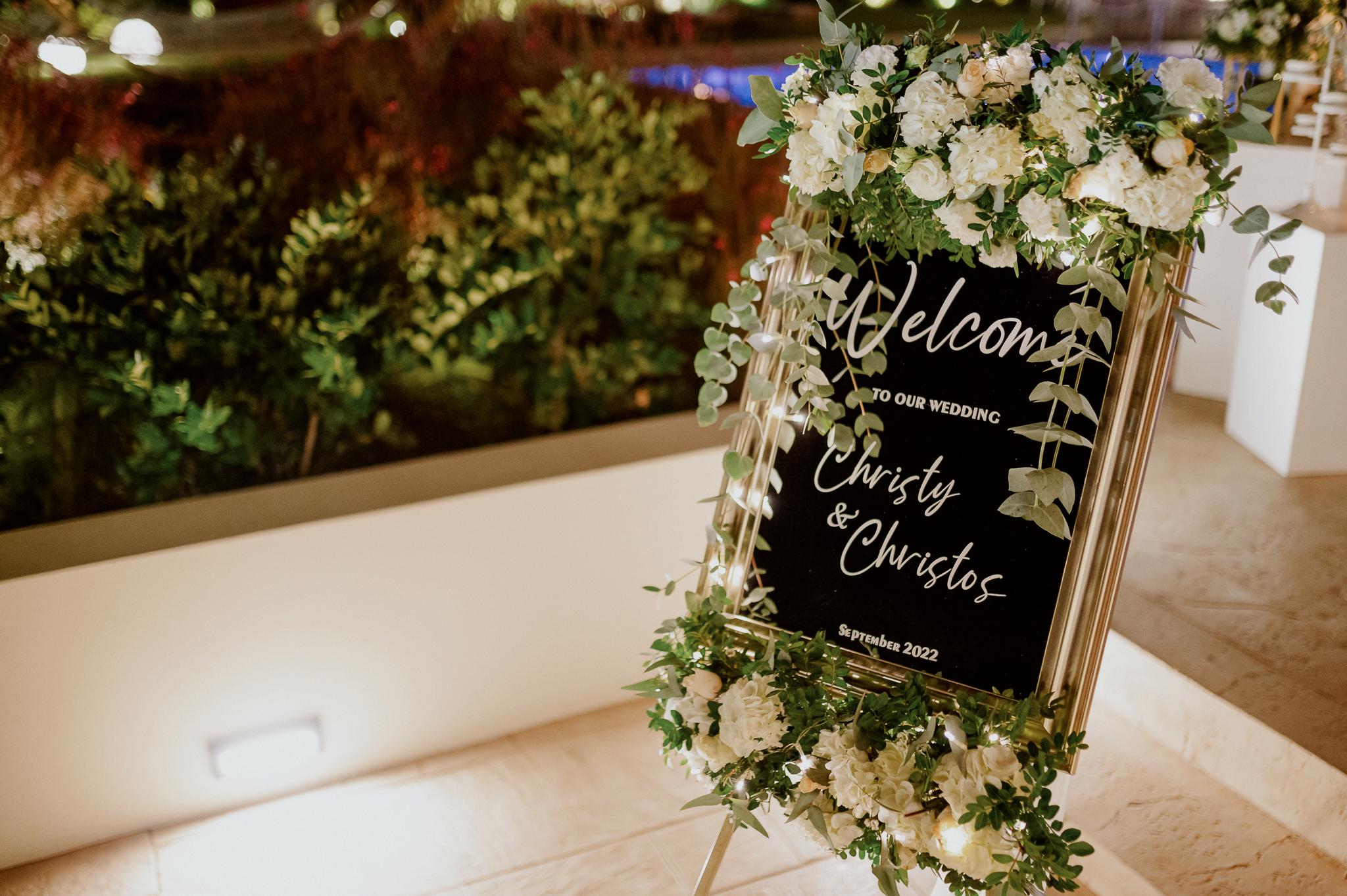 Κοντινή φωτογραφία από την σήμανση που βρίσκετε στην είσοδο του κτήματος η οποία αναγράφει καλώς ήρθατε στο γάμο μας και το όνομα του ζευγαριού . Επάνω στην σήμανση αυτή βρίσκετε μια σύνθεση με λουλούδια τριαντάφυλλα λευκά με πρασινάδες.