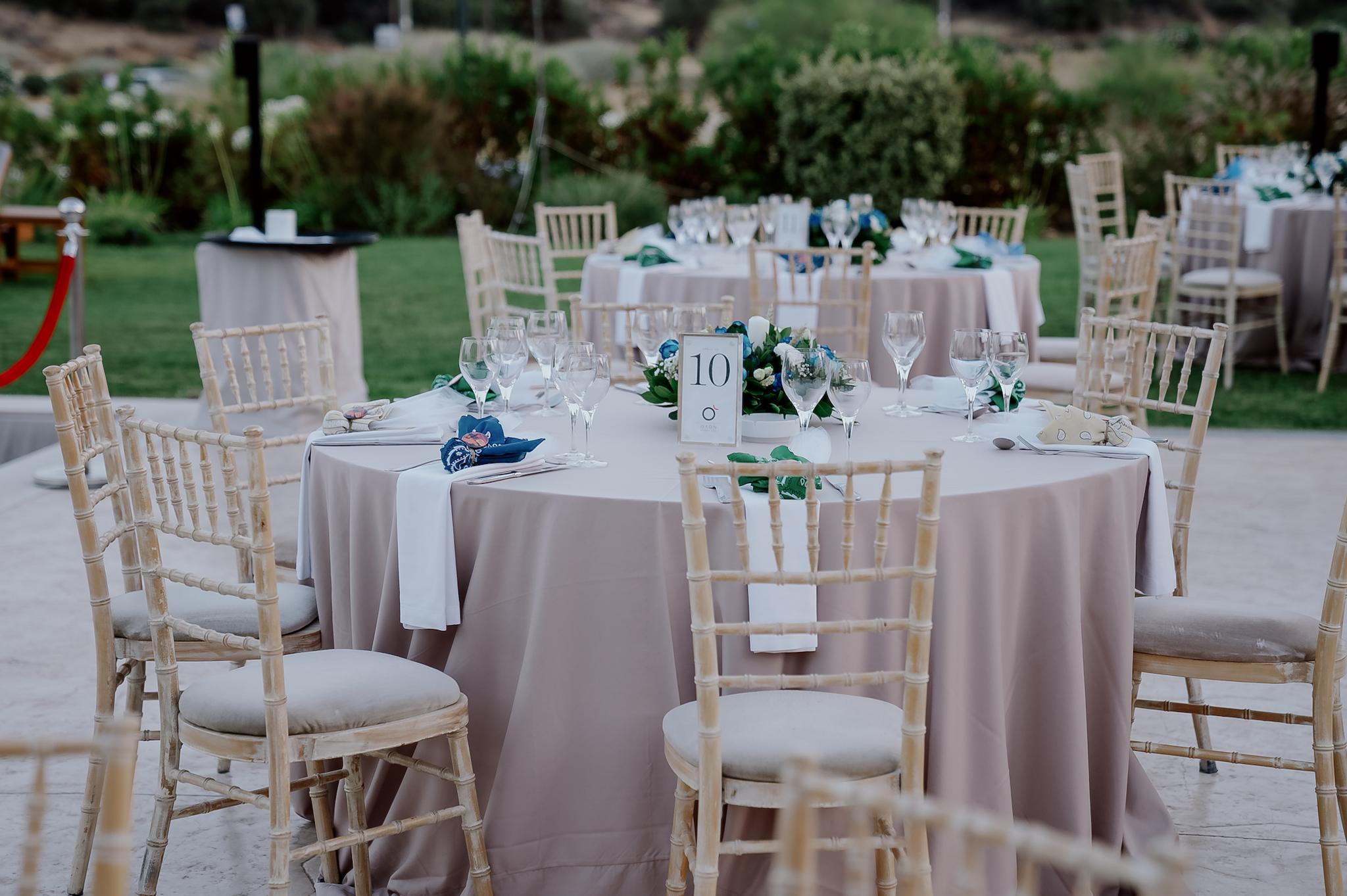 Μακρινή φωτογραφία στα τραπέζια του κτήματος , Λουλούδια βρίσκονται επάνω στα τραπέζια τα οποία περιέχουν μπλε τριαντάφυλλα και λευκά . Επάνω στο κάθε τραπέζι βρίσκονται τοποθετημένες και η μπομπονιέρες έχει στήσει το catering. Περιμετρικά από τις ροτόντες βρίσκονται οι tiffany ξύλινες καρέκλες