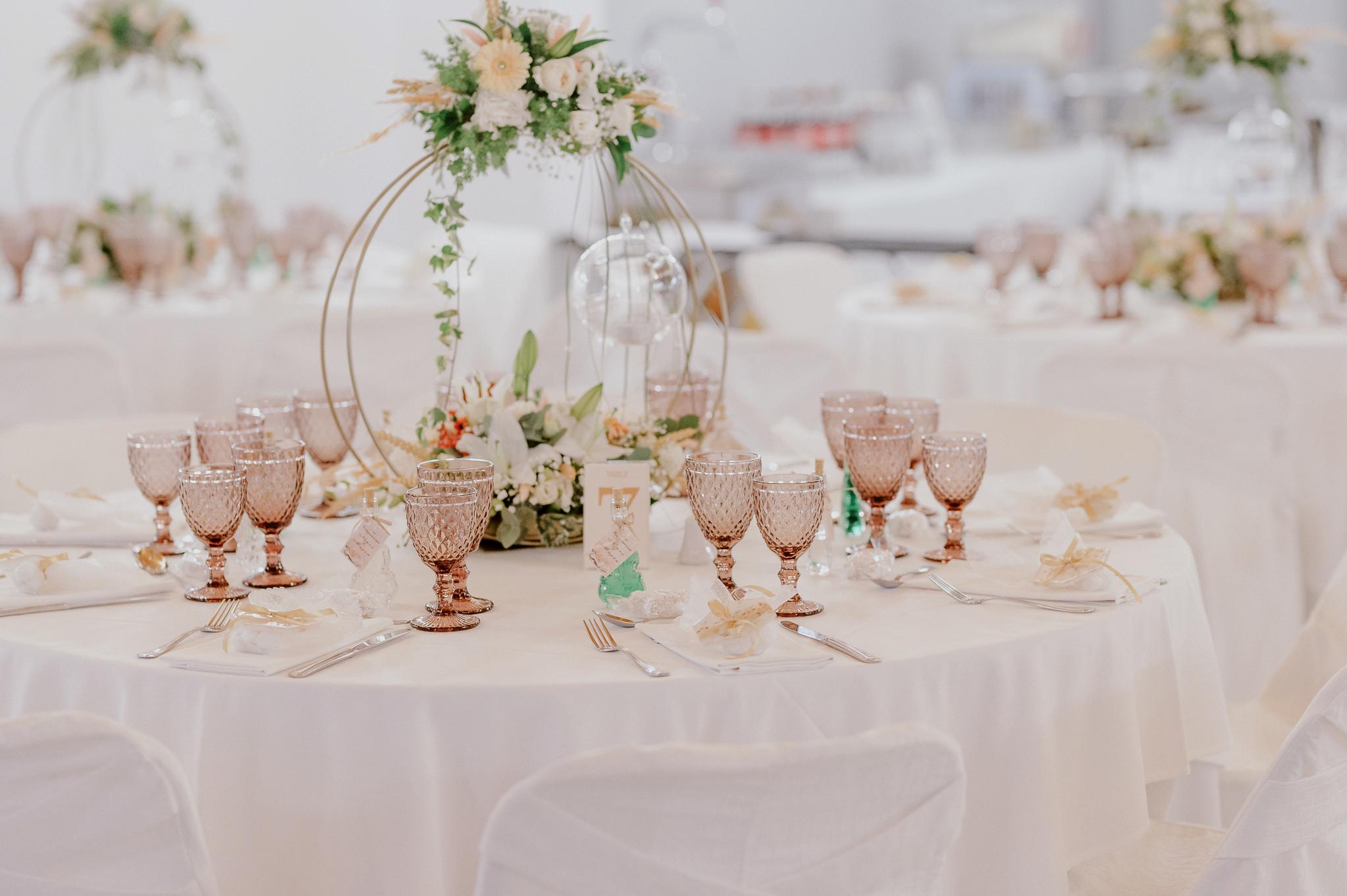 Φωτογραφία ροτόντας στον εσωτερικό χώρο του κτήματος με στολισμό του τραπεζιού με κερί να κρέμεται και στολισμό από ορχιδέα και τριαντάφυλλα σομόν με πράσινα φυτά. Στο τραπέζι βρίσκονται ποτήρια χρώματος ροζ και μαχαιροπίρουνα μαζί με συνοδευτικά γλυκά