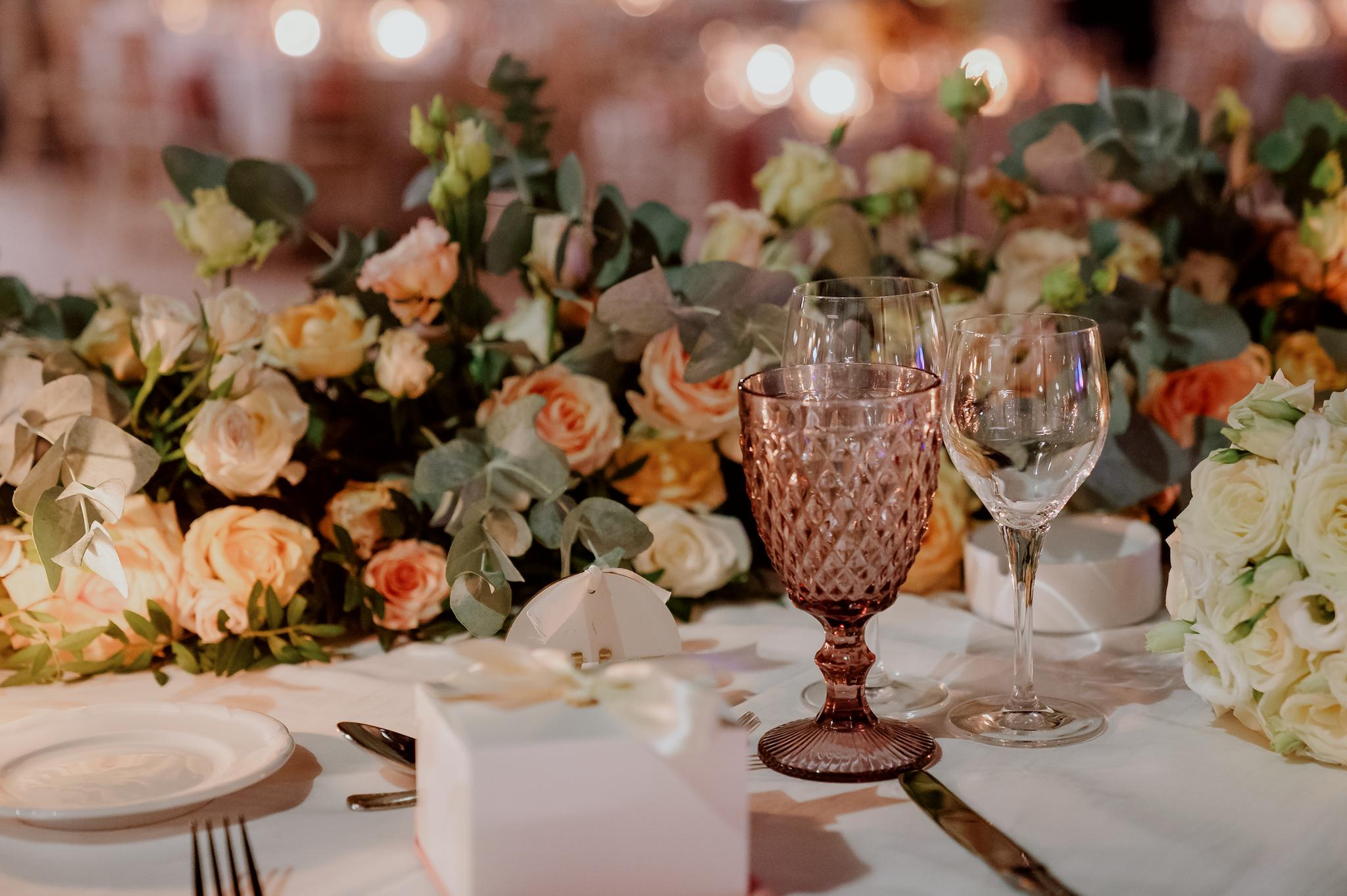 Κοντινή λήψη φωτογραφίας που απεικονίζει μπομπονιέρες επάνω στο τραπέζι στο κάθε άτομο ποτήρια από το catering μαχαιροπίρουνα στρωμένα τραπεζομάντηλα σε σομόν χρώμα