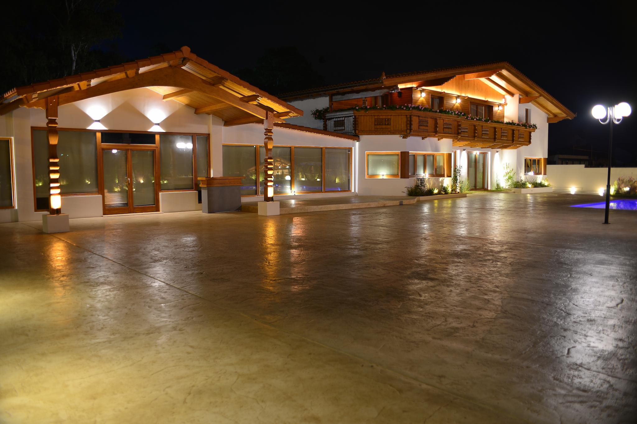 Φωτογραφία νυχτερινή η οποία δείχνει το ξύλινο ελβετικό σαλέ φωτισμένο. Στο βάθος φαίνεται και η πισίνα όπως επίσης και τον χώρο που τοποθετούνται τα τραπέζια.