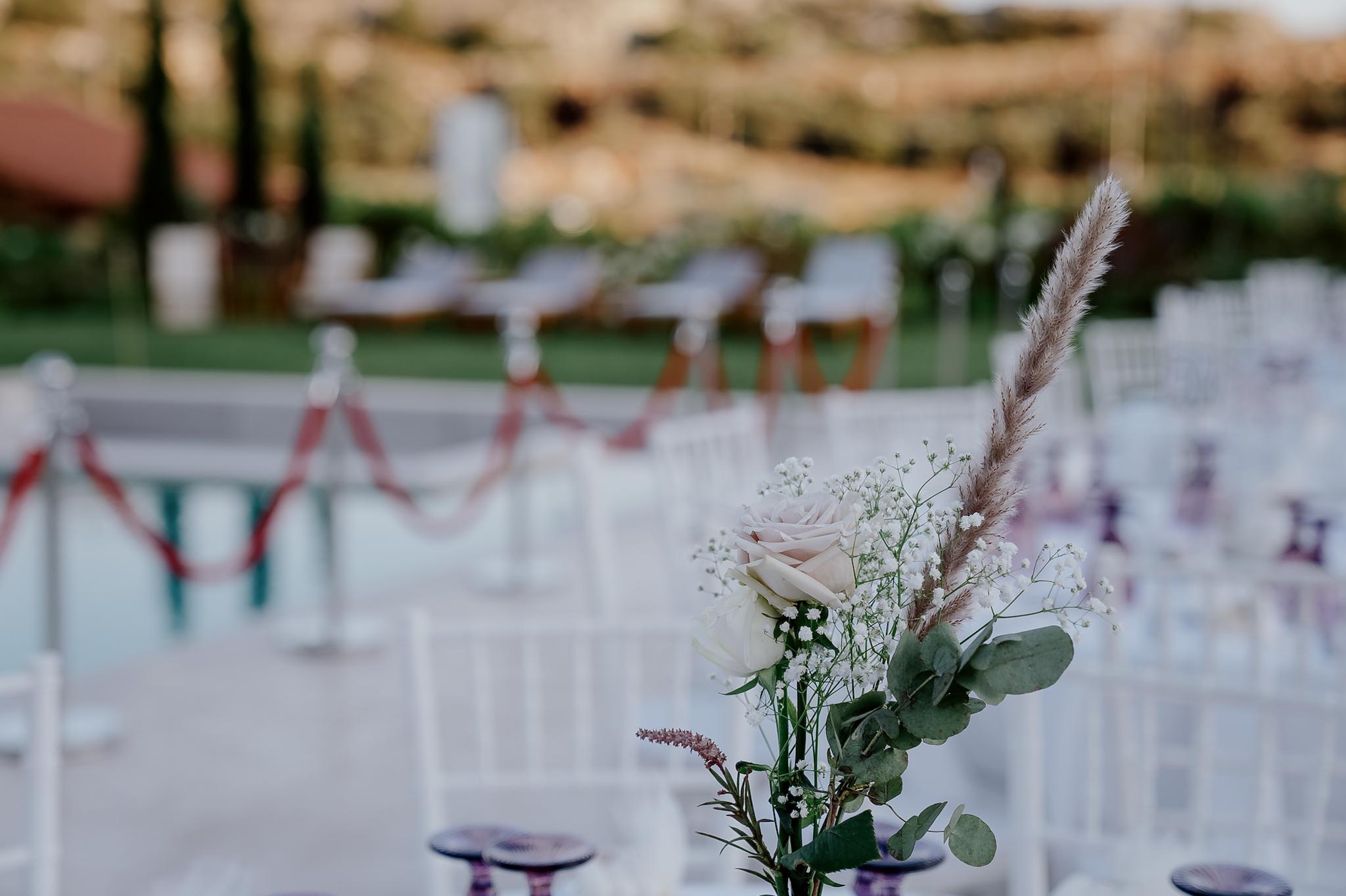 Κοντινή φωτογραφία που φαίνεται ο στολισμός του τραπεζιού ο οποίος είναι ένα σομόν τριαντάφυλλο με Pampas και πρασινάδα. Στο βάθος τις φωτογραφίας φαίνεται η πισίνας όπως επίσης και το πράσινο τοπίο που επικρατεί