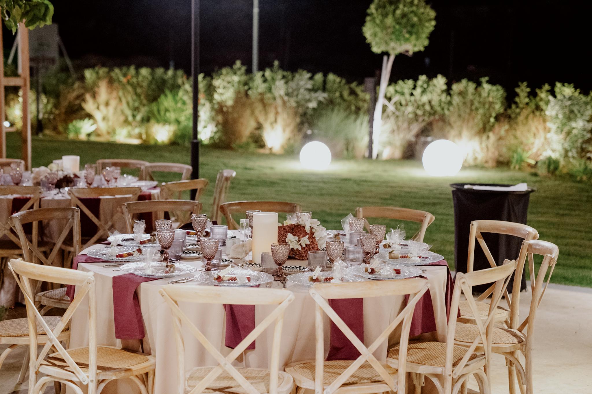 Κοντινή φωτογραφία με ροτόντες τραπέζια περιμετρικά από τα τραπέζια είναι ξύλινες καρέκλες. Τα τραπέζια έχουν μπεζ τραπεζομάντηλα και επάνω μπορντό ατομικές πετσέτες. Στο βάθος είναι το αυτοκινούμενο βαν μπαρ το οποίο το χρησιμοποιούν οι καλεσμένοι για να παίρνουν ποτό . Στο βάθος φαίνεται το γκαζόν αλλά και τα φυτά που έχει το κτήμα.