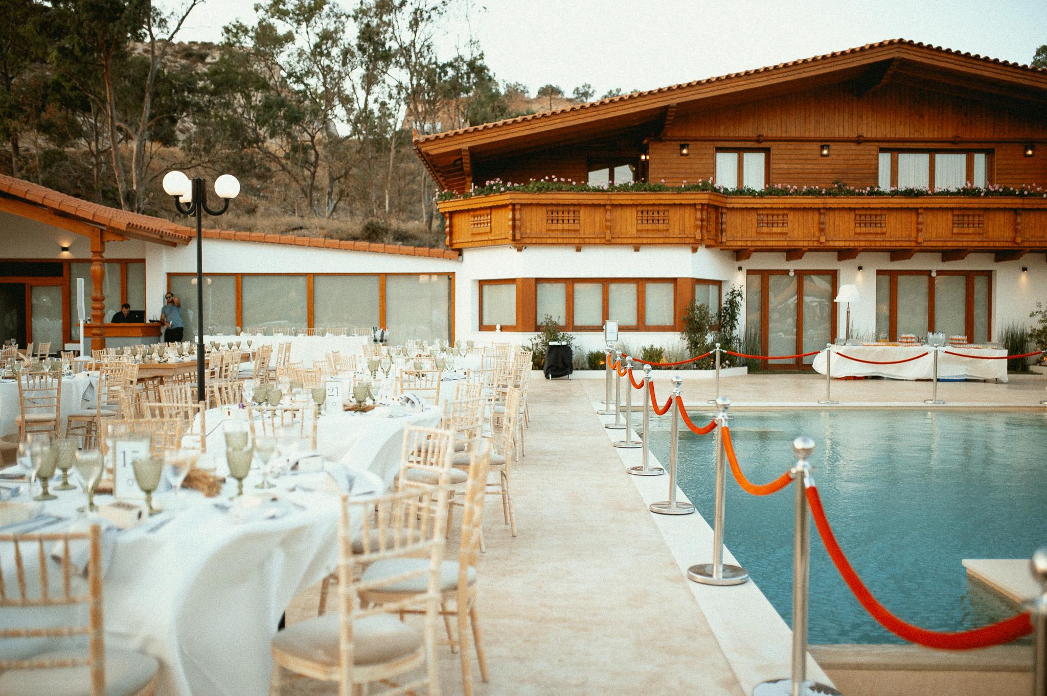 Απομακρυσμένη φωτογραφία πρωινή με στρωμένες ροτόντες και μοναστηριακά τραπέζια. Περιμετρικά από τα τραπέζια είναι στημένες οι καρέκλες ξύλινες tiffany. Στο βάθος τις φωτογραφίας φαίνεται το ξύλινο σαλέ που υπάρχει εντός του κτήματος . Στην μισή φωτογραφία φαίνεται η πισίνα