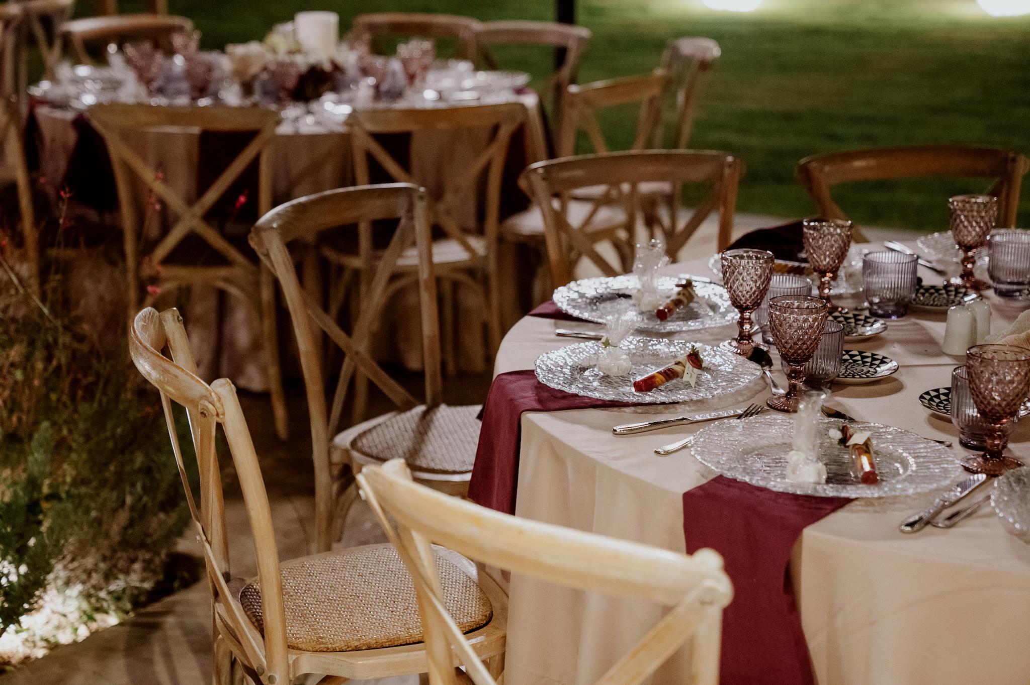 Κοντινή φωτογραφία με ροτόντες τραπέζια περιμετρικά από τα τραπέζια είναι ξύλινες καρέκλες. Τα τραπέζια έχουν μπεζ τραπεζομάντηλα και επάνω μπορντό ατομικές πετσέτες. Στο βάθος είναι το αυτοκινούμενο βαν μπαρ το οποίο το χρησιμοποιούν οι καλεσμένοι για να παίρνουν ποτό .