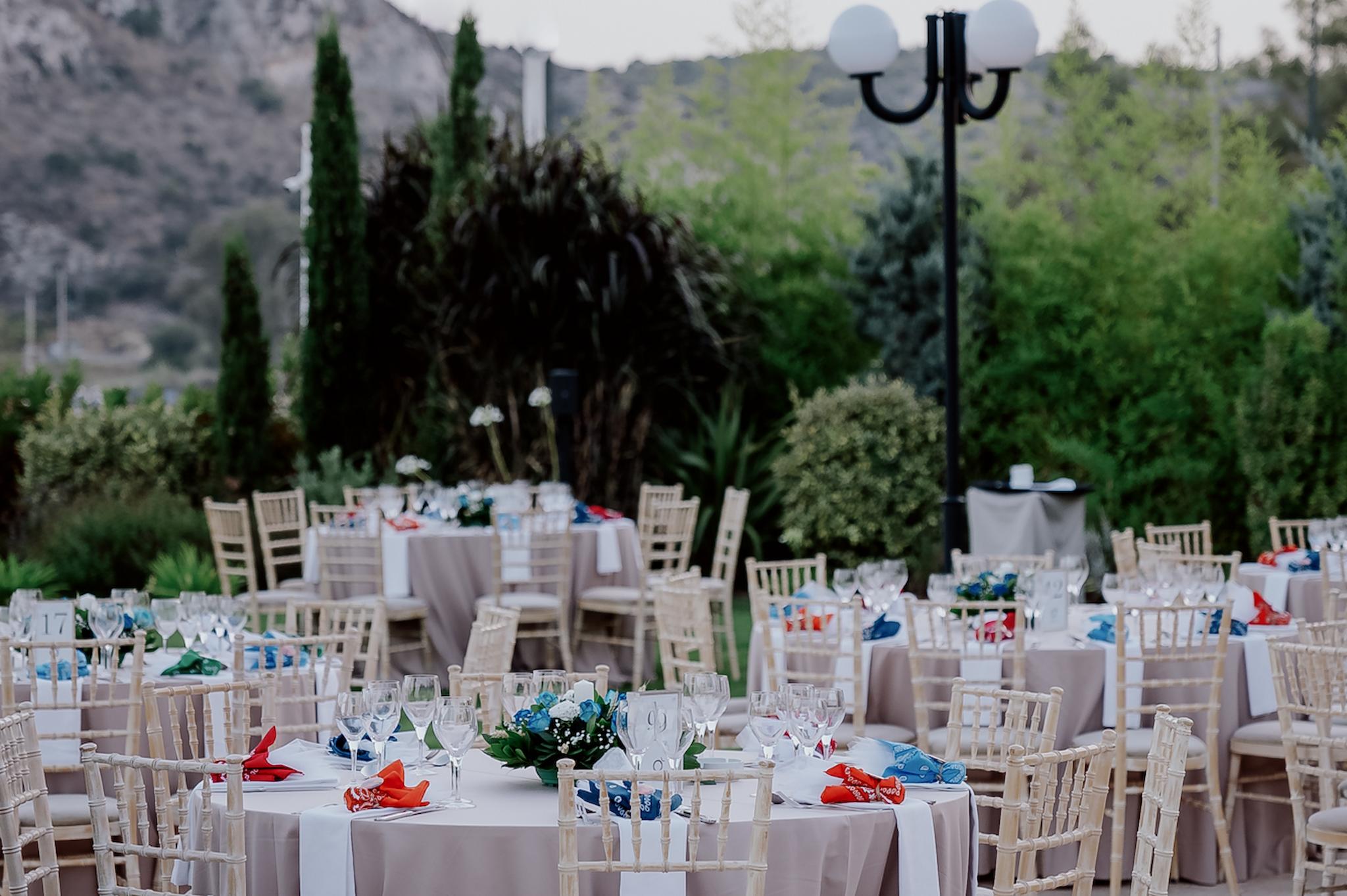 Μακρινή φωτογραφία στα τραπέζια του κτήματος , λουλούδια βρίσκονται επάνω στα τραπέζια τα οποία περιέχουν μπλε τριαντάφυλλα και λευκά . Επάνω στο κάθε τραπέζι βρίσκονται τοποθετημένες και η μπομπονιέρες. Στο βάθος φαίνεται ο πράσινος κήπος του κτήματος