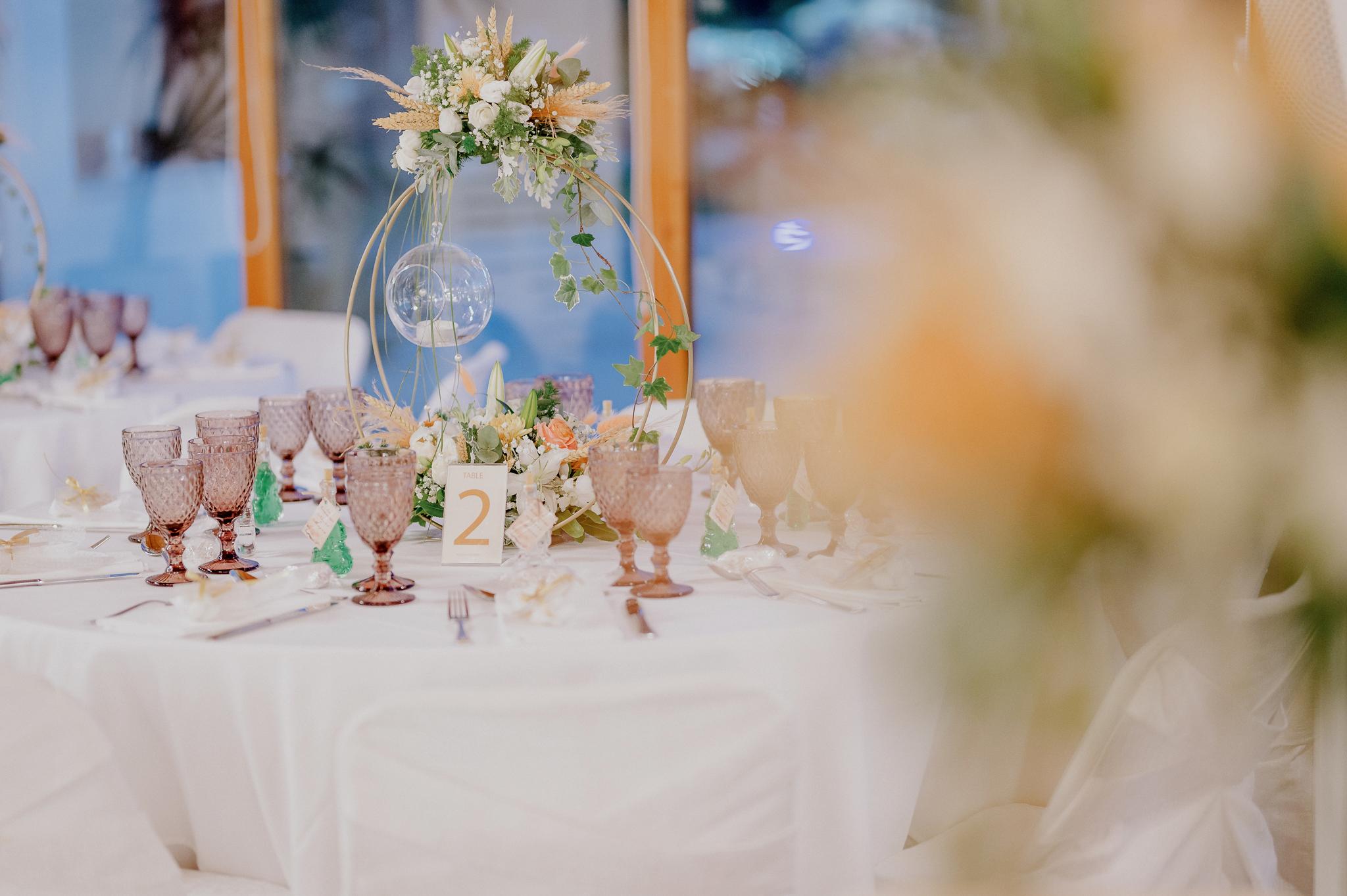 Φωτογραφία ροτόντας στον εσωτερικό χώρο του κτήματος με στολισμό του τραπεζιού με κερί να κρέμεται και στολισμό από ορχιδέα και τριαντάφυλλα σομόν με πράσινα φυτά. Στο τραπέζι βρίσκονται ποτήρια χρώματος ροζ και μαχαιροπίρουνα μαζί με συνοδευτικά γλυκά. Στο βάθος φαίνεται η πισίνα στον εξωτερικό χώρο από τις τζαμαρίες που έχει ο εσωτερικός χώρος μας .